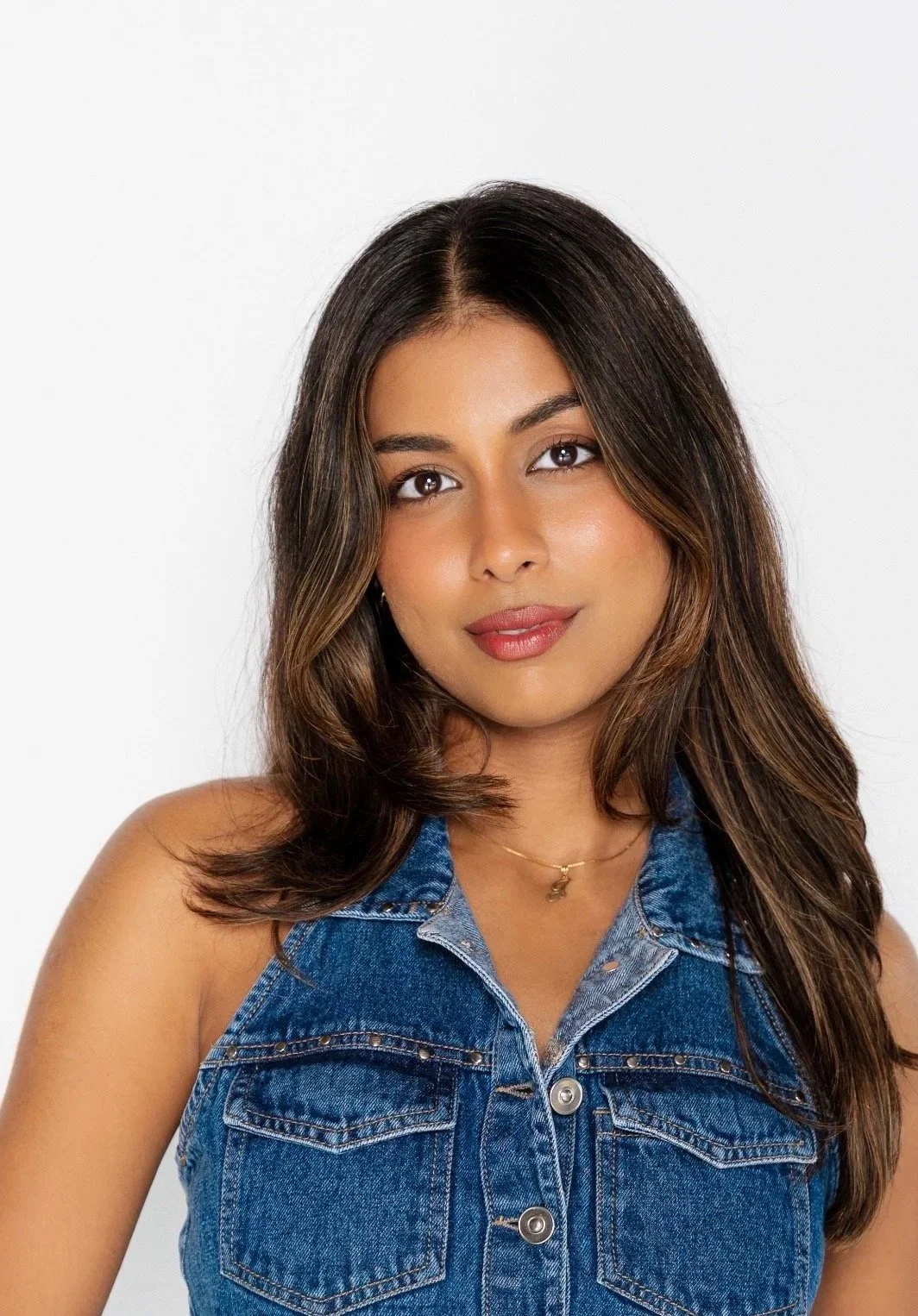 A young woman with long brown hair, wearing a denim sleeveless shirt, and a delicate gold necklace with a small pendant, posing against a plain white background.