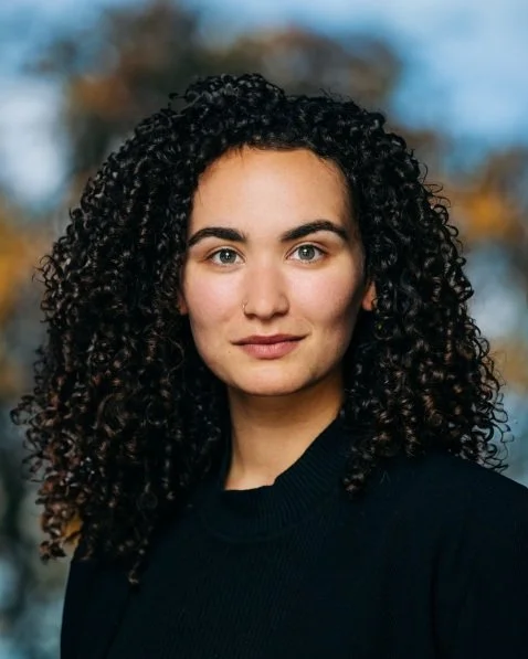 A young woman with curly dark hair and light skin, outdoors with blurred trees in the background.