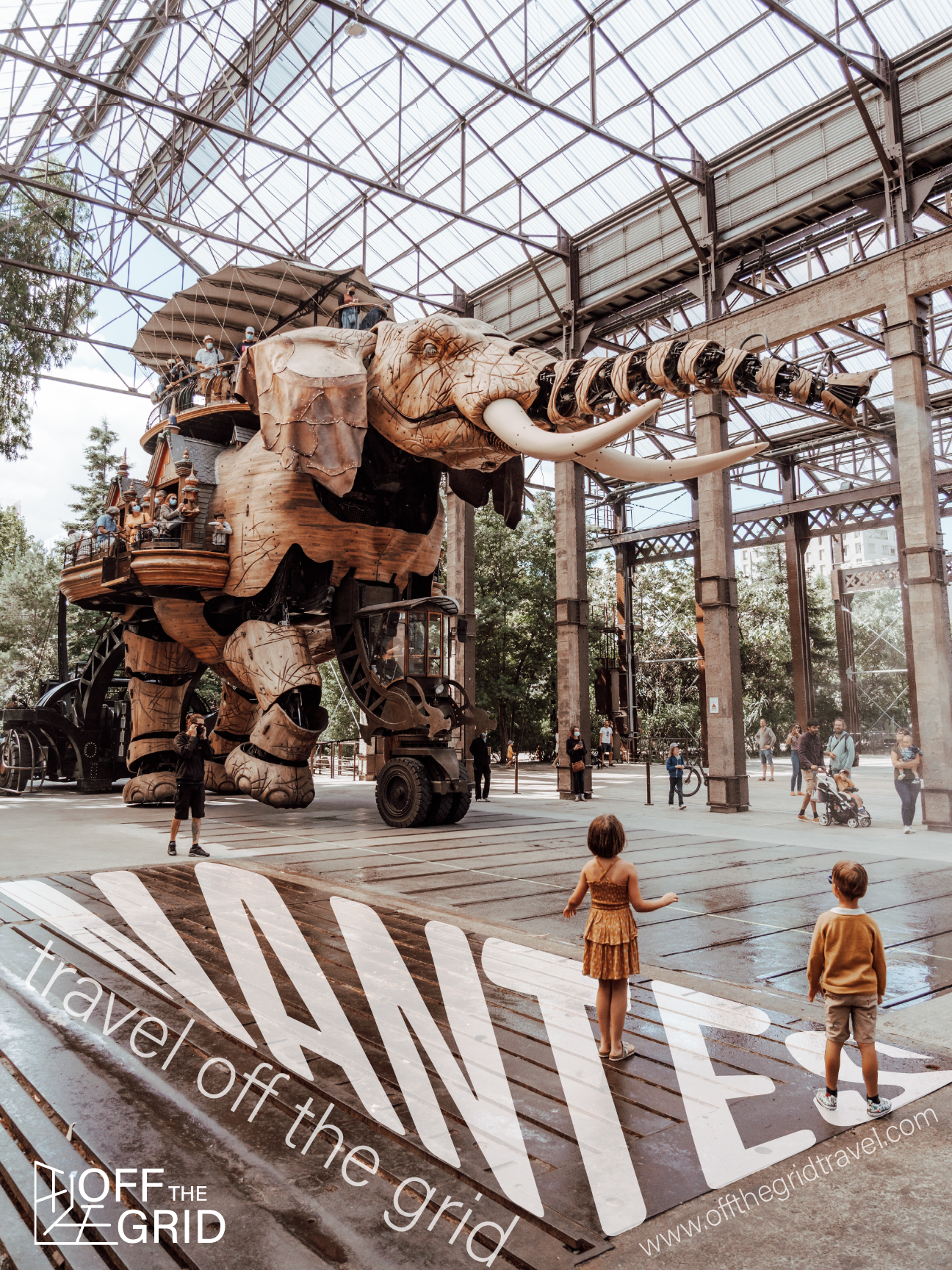 A large, elephant-shaped mechanical sculpture with people riding on and around it, located inside a spacious industrial-style space with a glass ceiling, with children and adults observing and walking around.