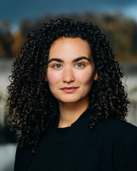 A young woman with curly dark hair and light skin, wearing a black sweater outdoors with a blurred background of trees and a cloudy sky.