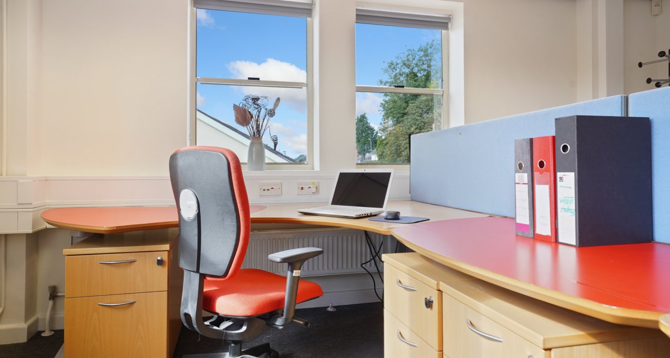 Small office space with a red chair tucked in beneath a curved desk that overlooks two windows.
