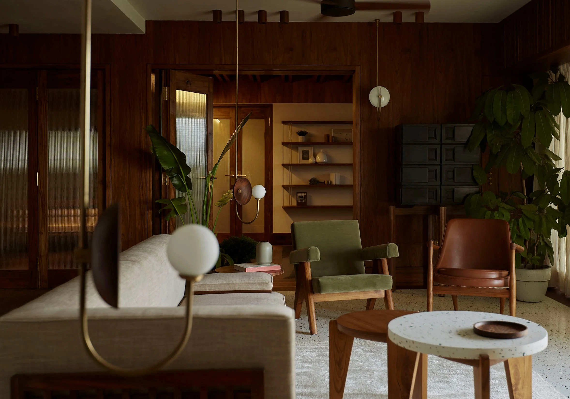 Mid-century modern living room with wooden decor, featuring a green armchair, leather chair, light gray sofa, round white speckled coffee table, indoor plants, pendant lighting, and open shelves. Warm lighting creates a cozy ambiance.