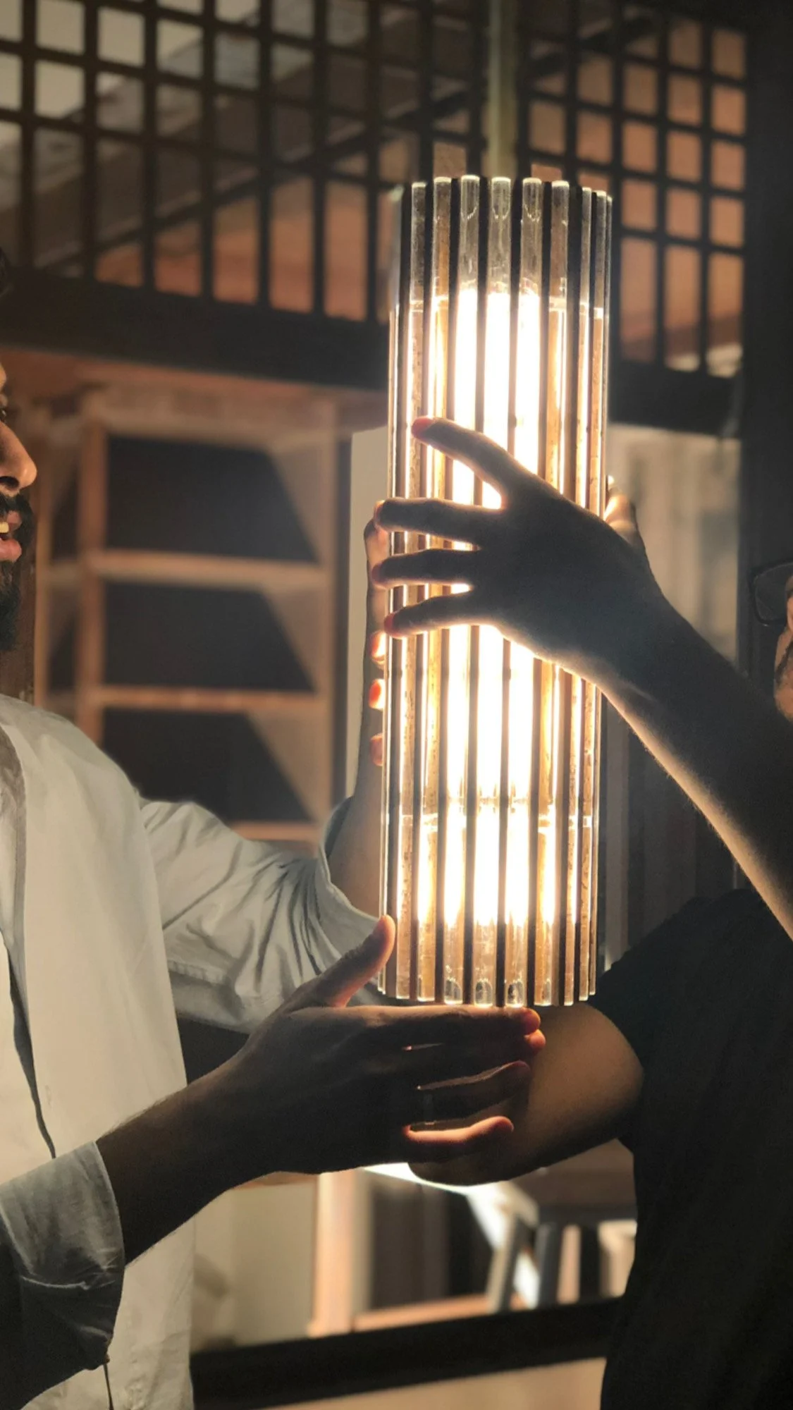 Two people holding a glowing cylindrical light fixture in a dimly lit room.