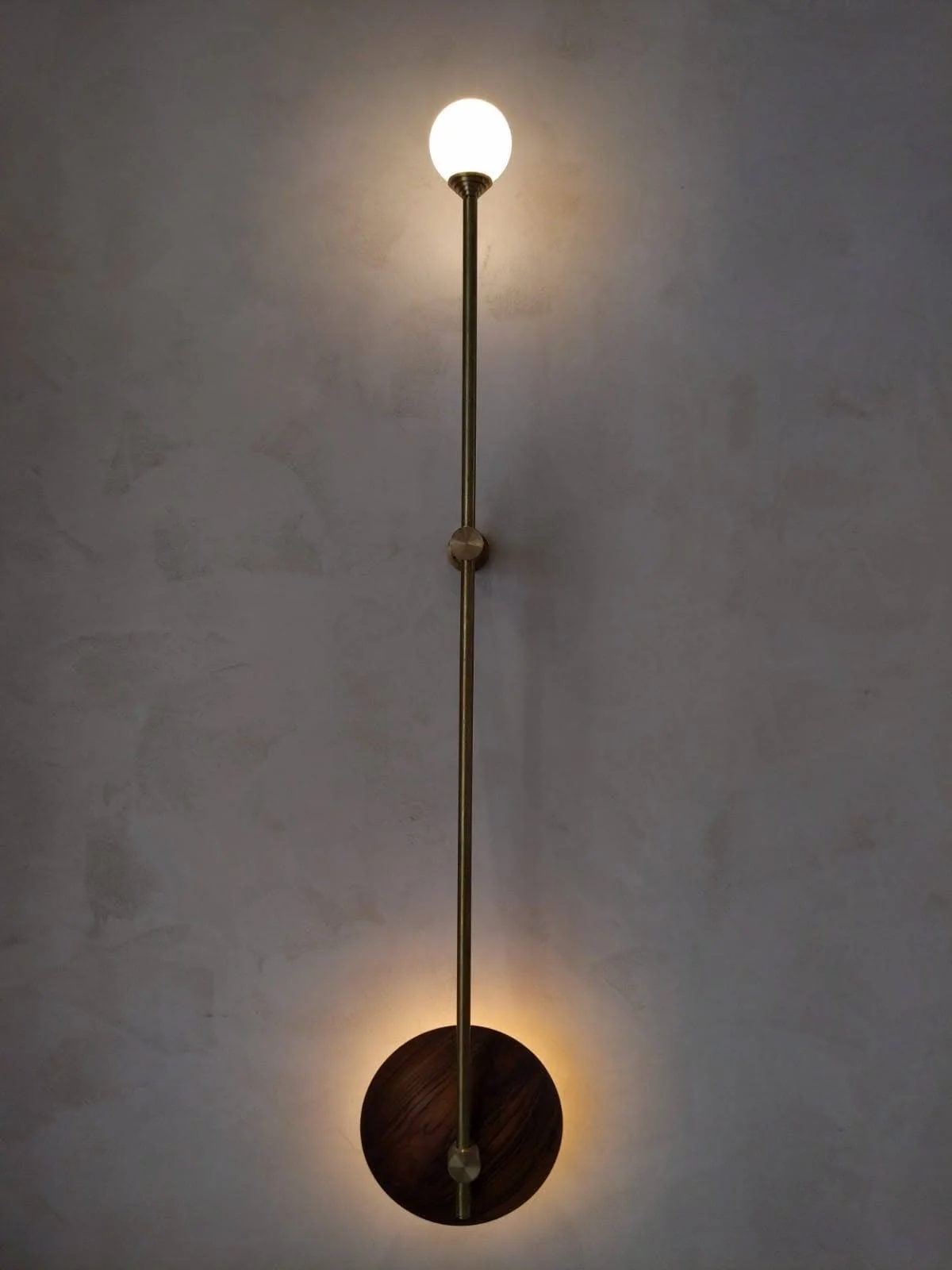 Vertical wall sconce with a glowing round bulb at the top and a circular light base at the bottom, mounted on a textured wall.