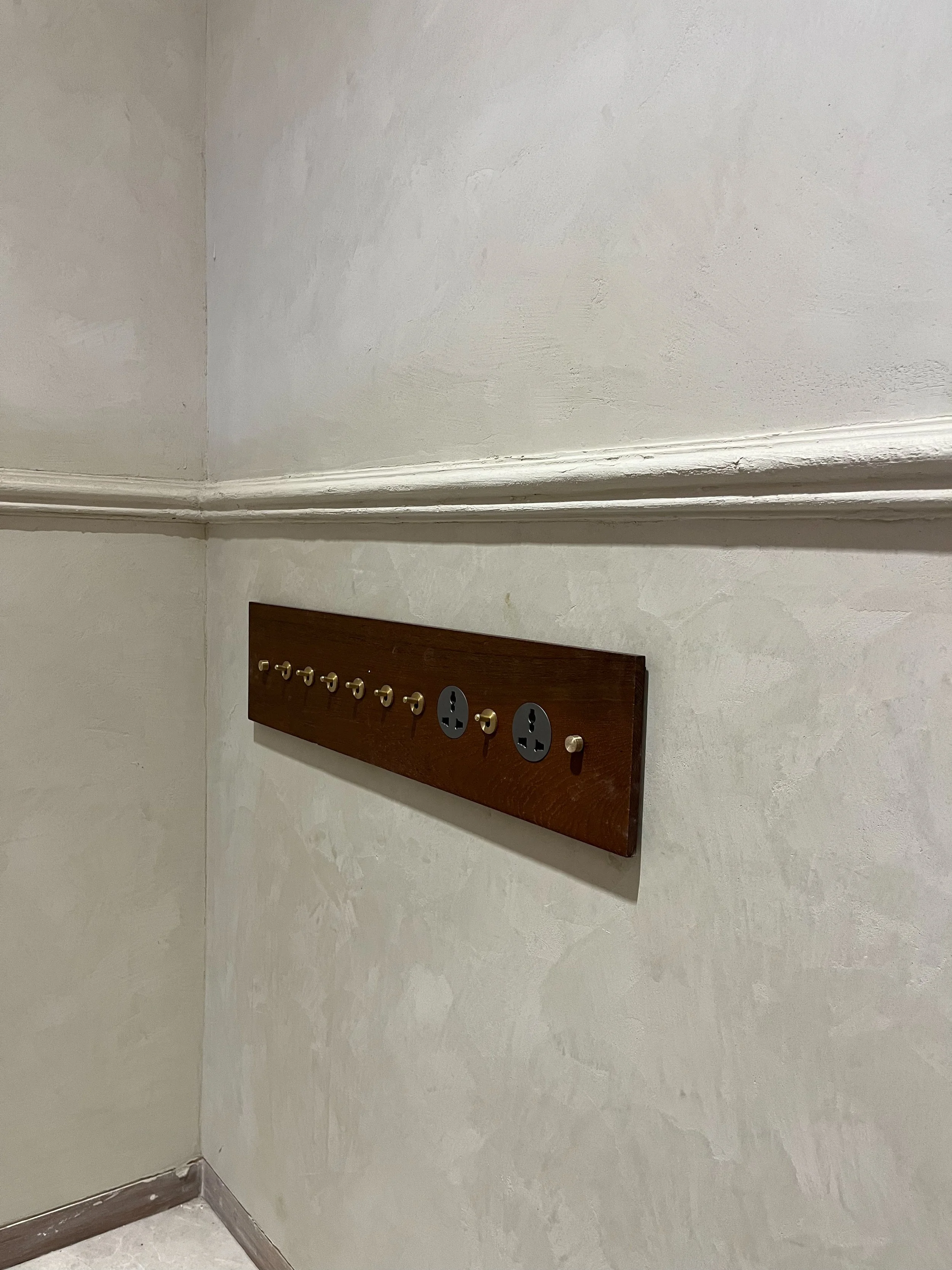 Vintage wall panel with multiple toggle switches and two electrical outlets, mounted on a painted wall corner.