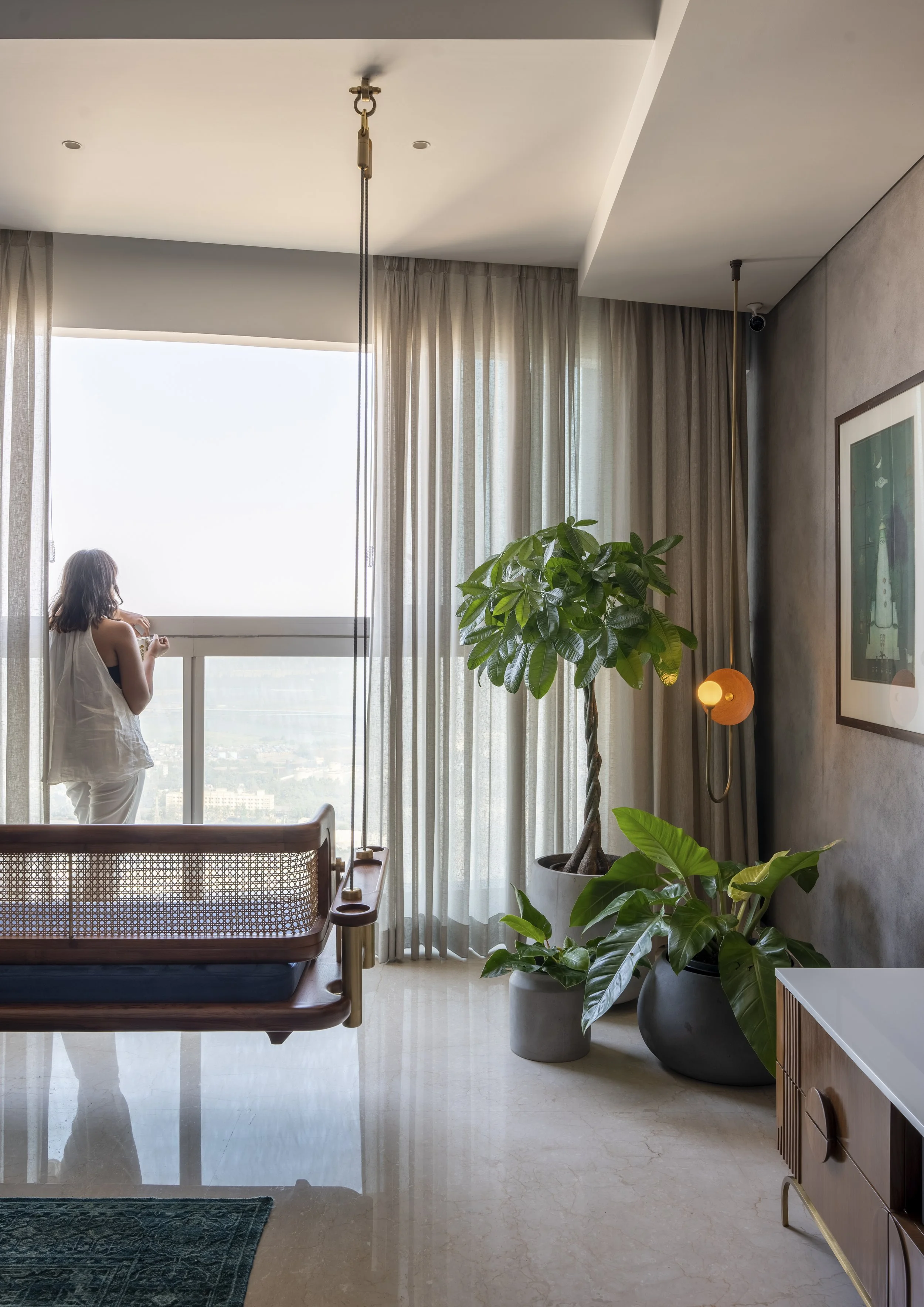Interior space with a woman standing by a large window, sheer curtains, and a modern hanging chair. Large potted plants decorate the room, which features a marble floor and a minimalist decor style.