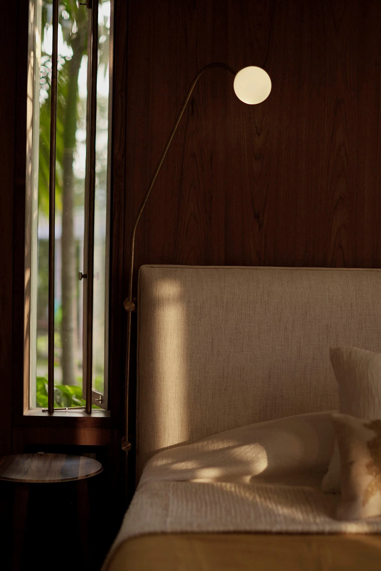 Interior of a cozy bedroom with partial view of a bed, wooden wall, nightstand, round lamp, and window showing greenery outside.