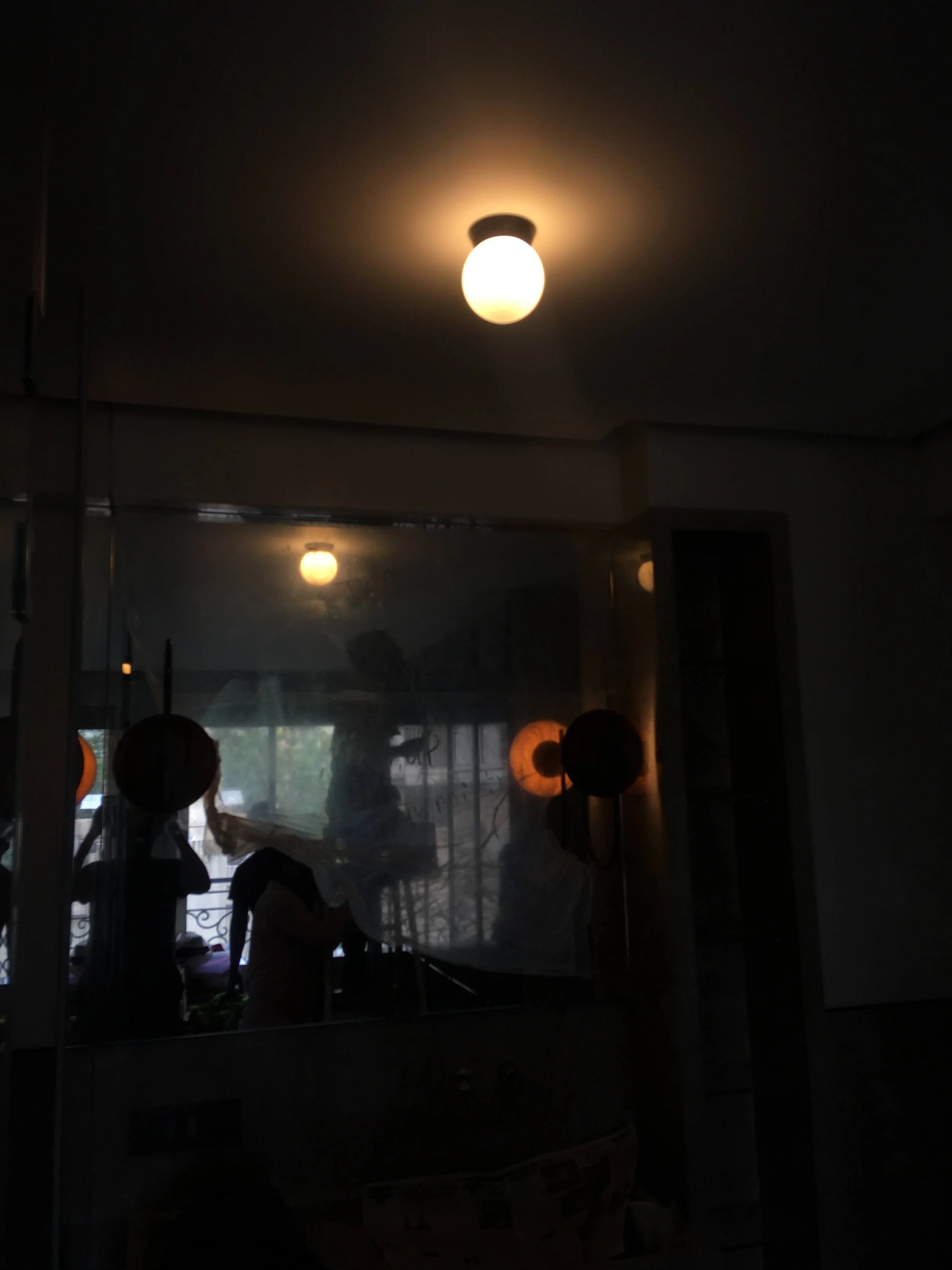 Dimly lit room with a ceiling light and reflections in a mirror, showing silhouettes and objects.