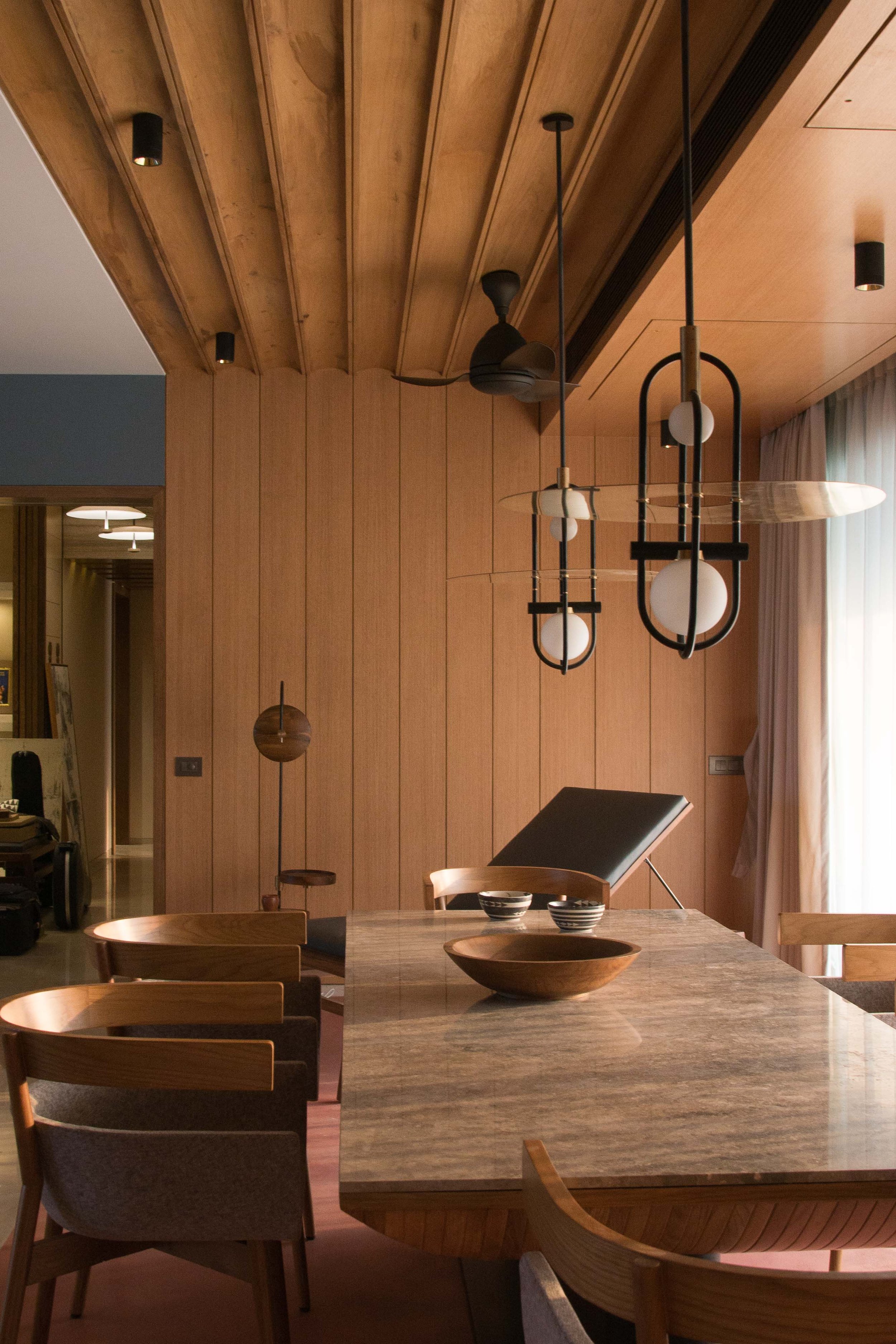 A modern dining room featuring a wooden table with chairs, a ceiling fan, pendant lights, and wooden panel walls. A decorative bowl and bowls are on the table, with a chaise lounge visible in the background.