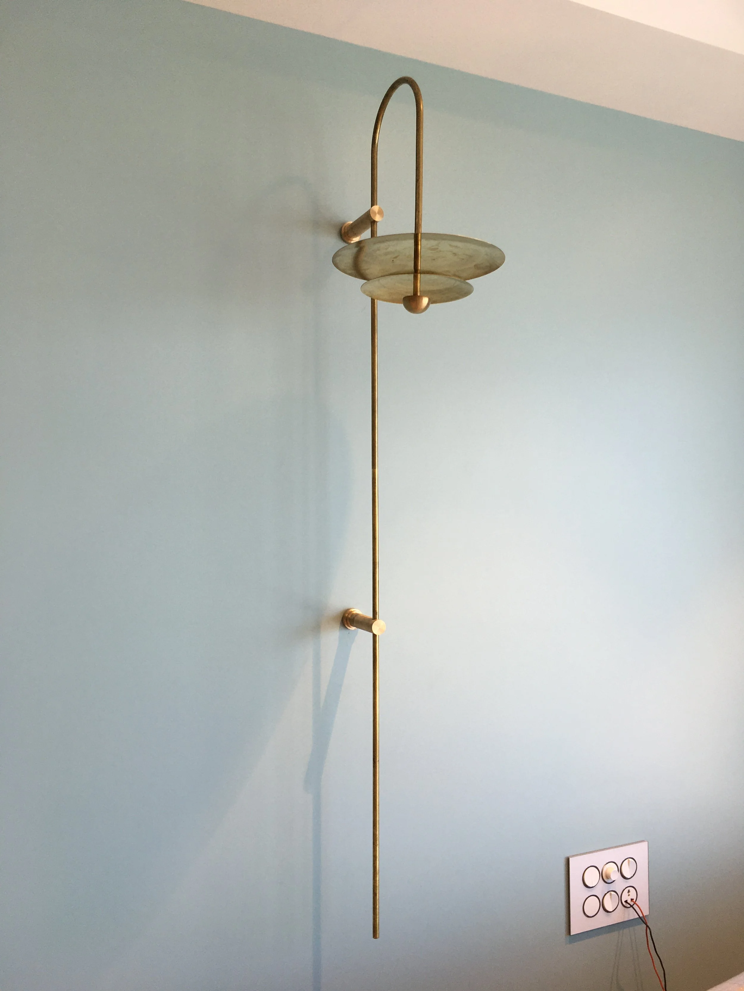 Brass wall-mounted lamp with a circular shade on a blue wall, featuring exposed screws and wiring near a light switch panel.