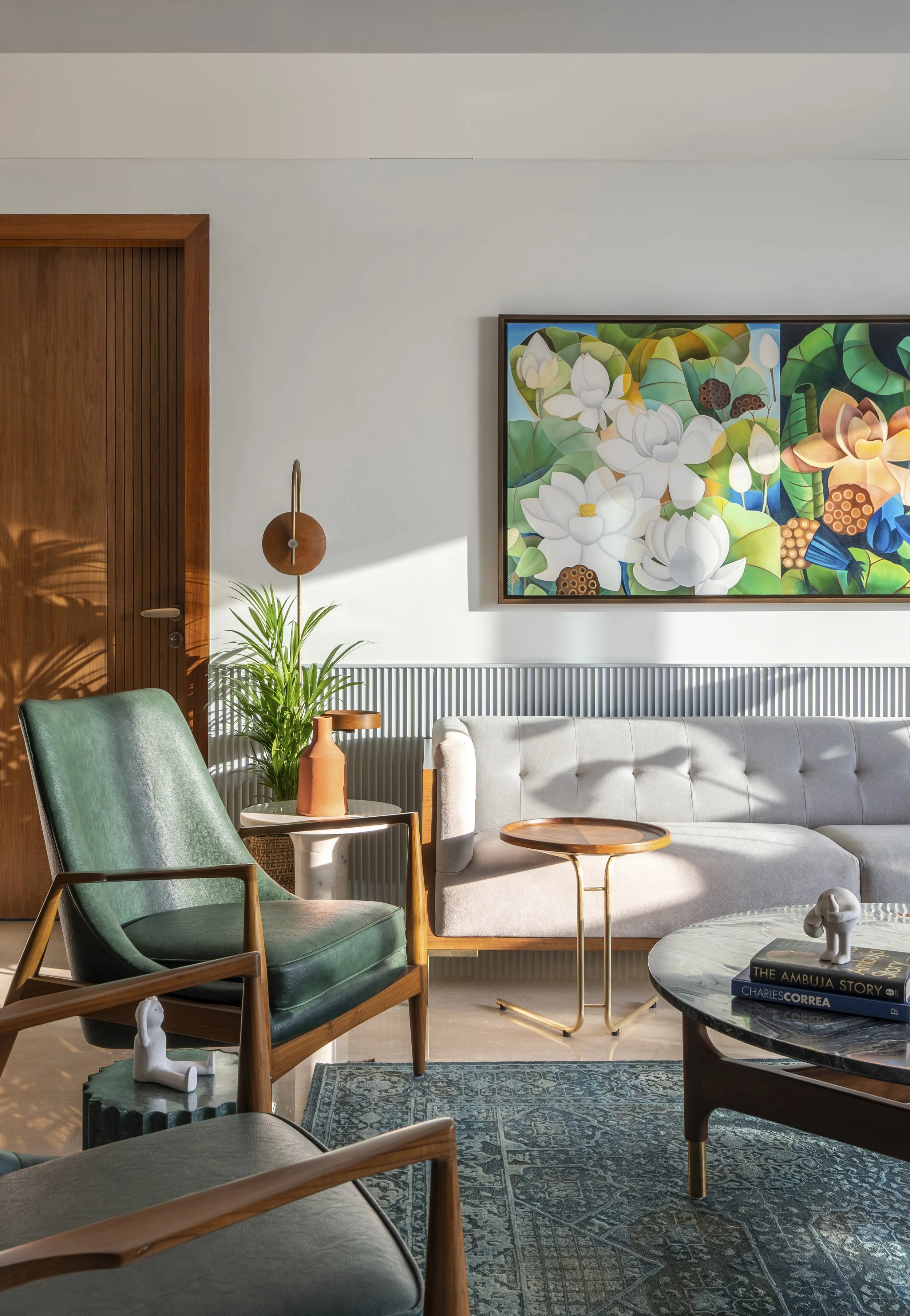 Stylish living room with a green leather armchair, gray tufted sofa, and round wooden side tables. A colorful floral painting hangs on the white wall, and there is a potted plant beside the wooden door. A patterned rug and books on a marble coffee ta
