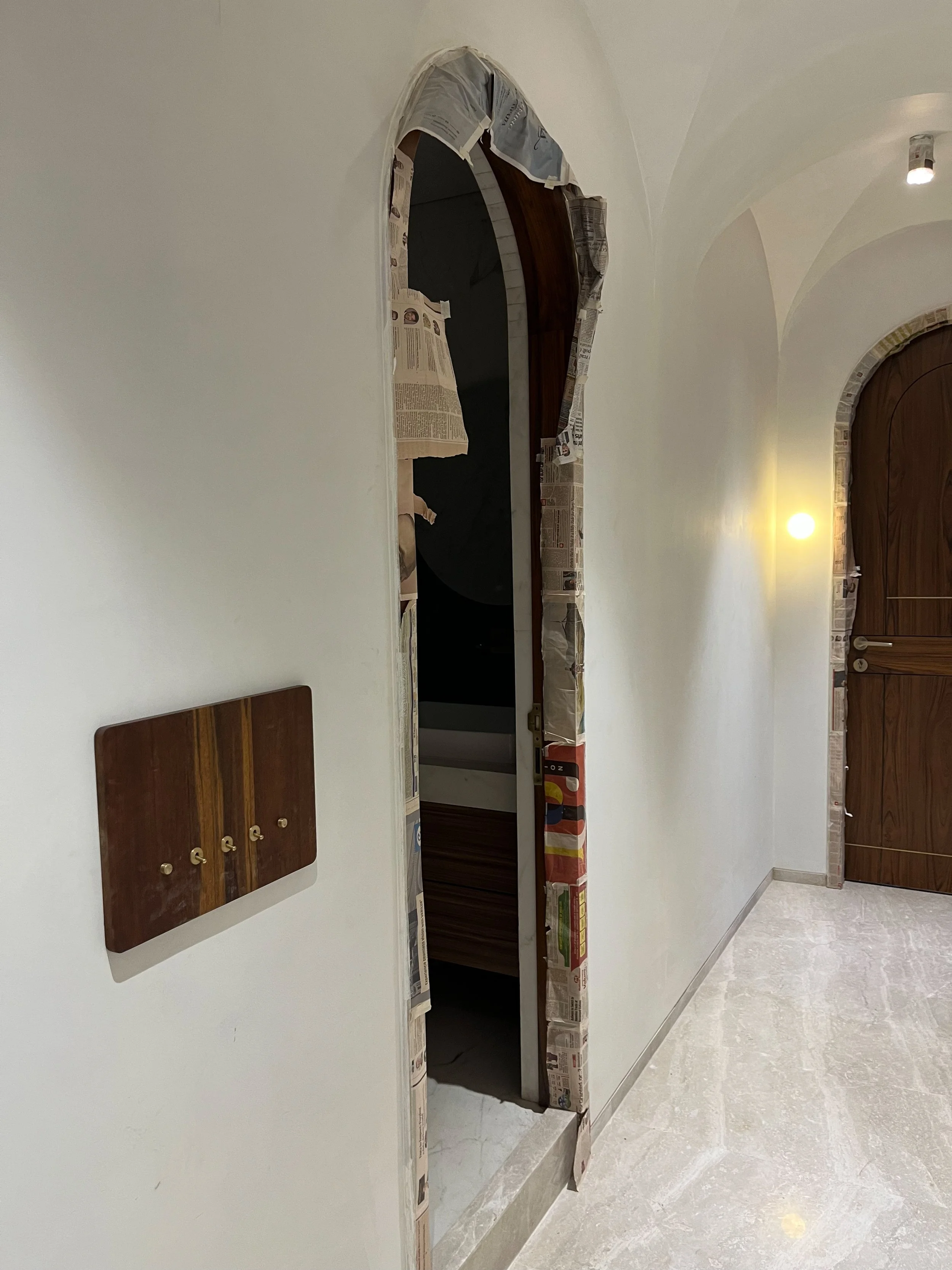Hallway with light colored walls and flooring, partially open wooden door with newspaper taped around the frame, adjacent closed wooden door, wall-mounted wooden panel.