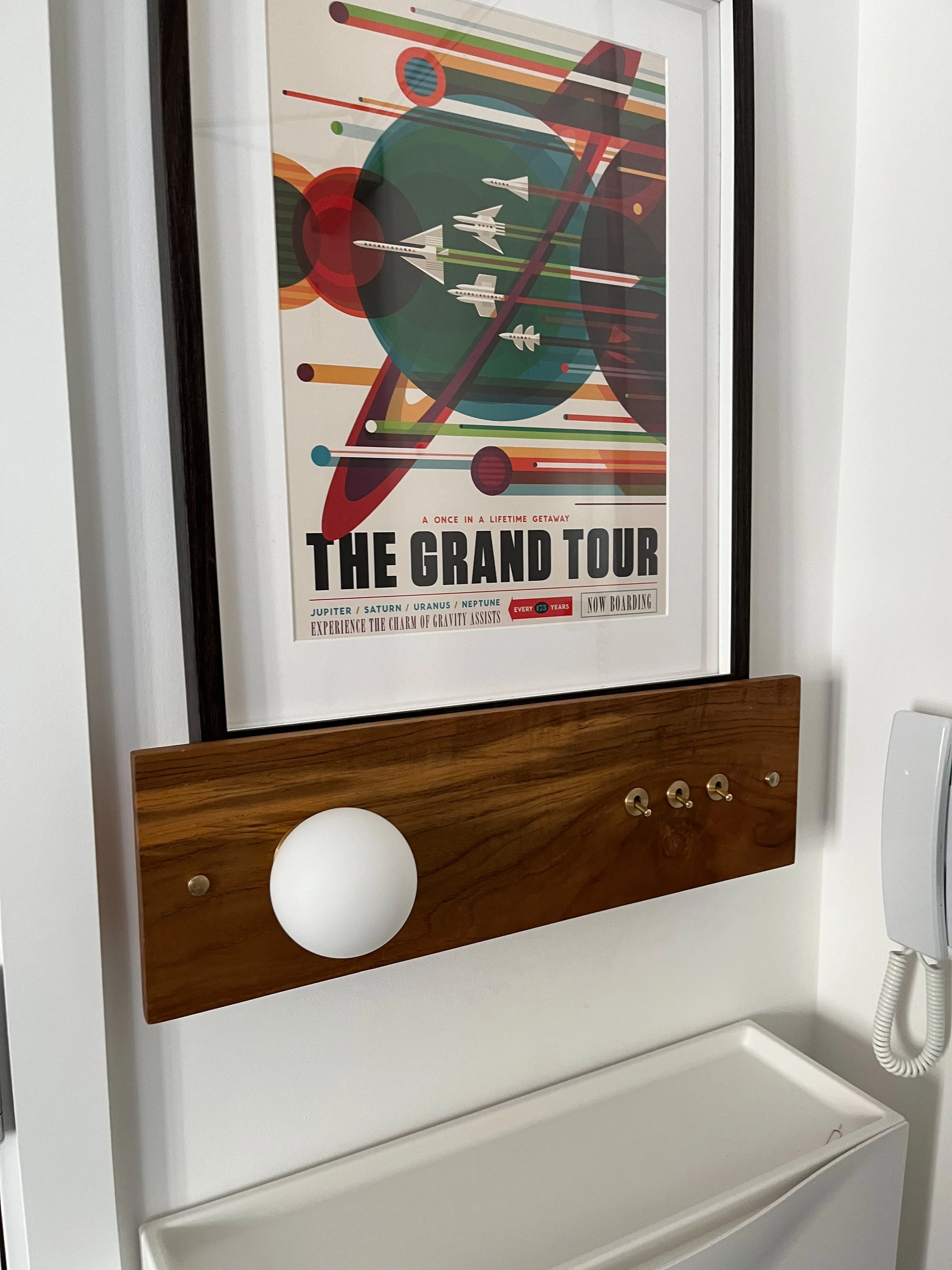Framed poster with retro space-themed art and text "The Grand Tour," mounted above a wooden light switch panel on a wall.
