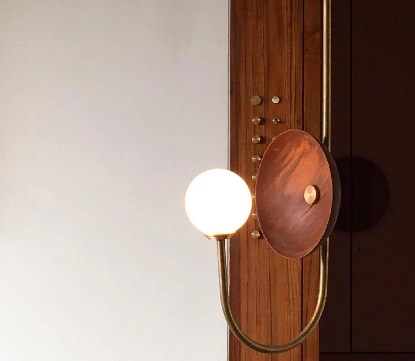 Modern wall lamp with a spherical light bulb and a wooden backplate, on a wood-paneled wall with circular brass accents.