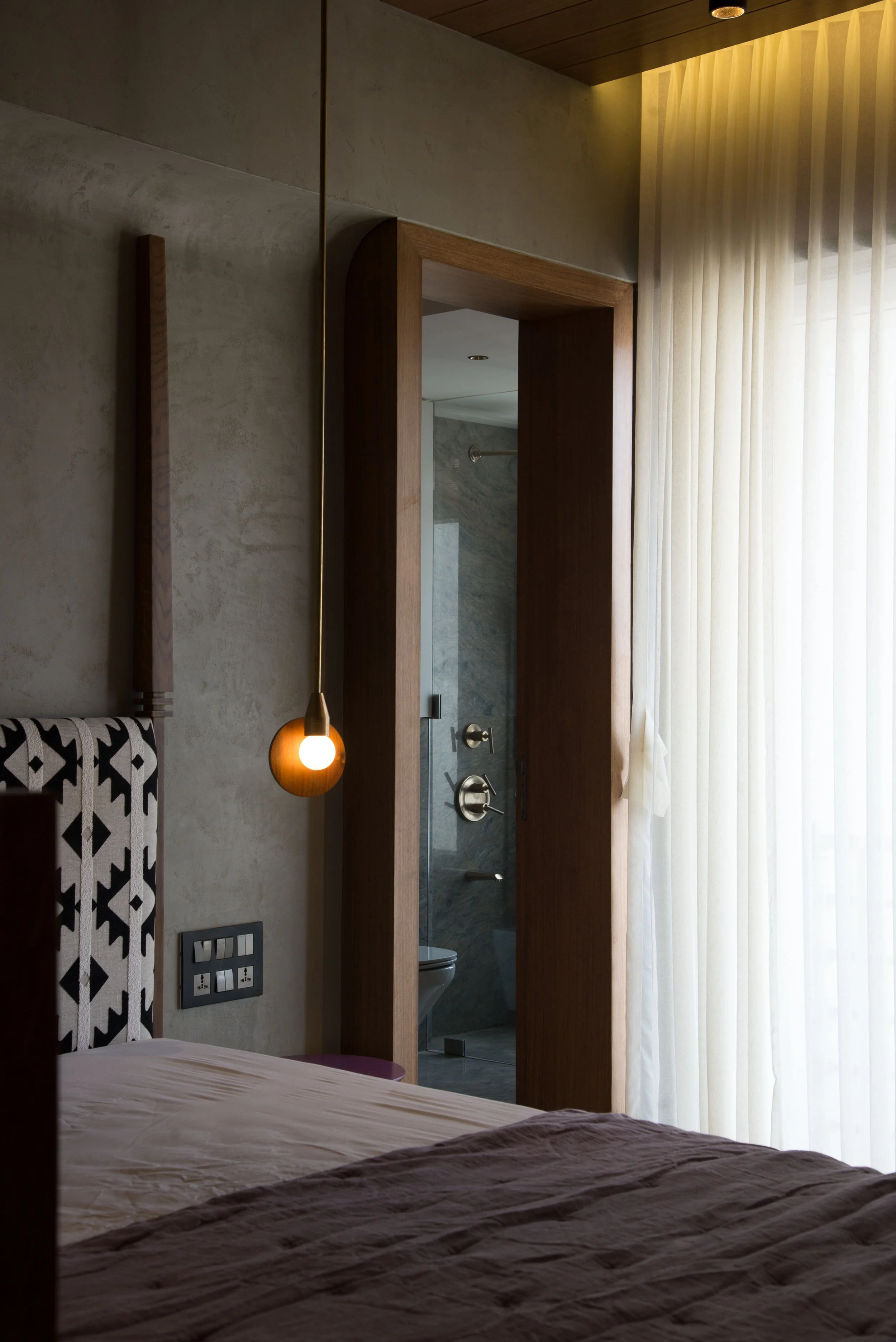Modern bedroom interior with hanging light, patterned headboard, wooden accents, and view into bathroom. Floor-to-ceiling curtain filters natural light.