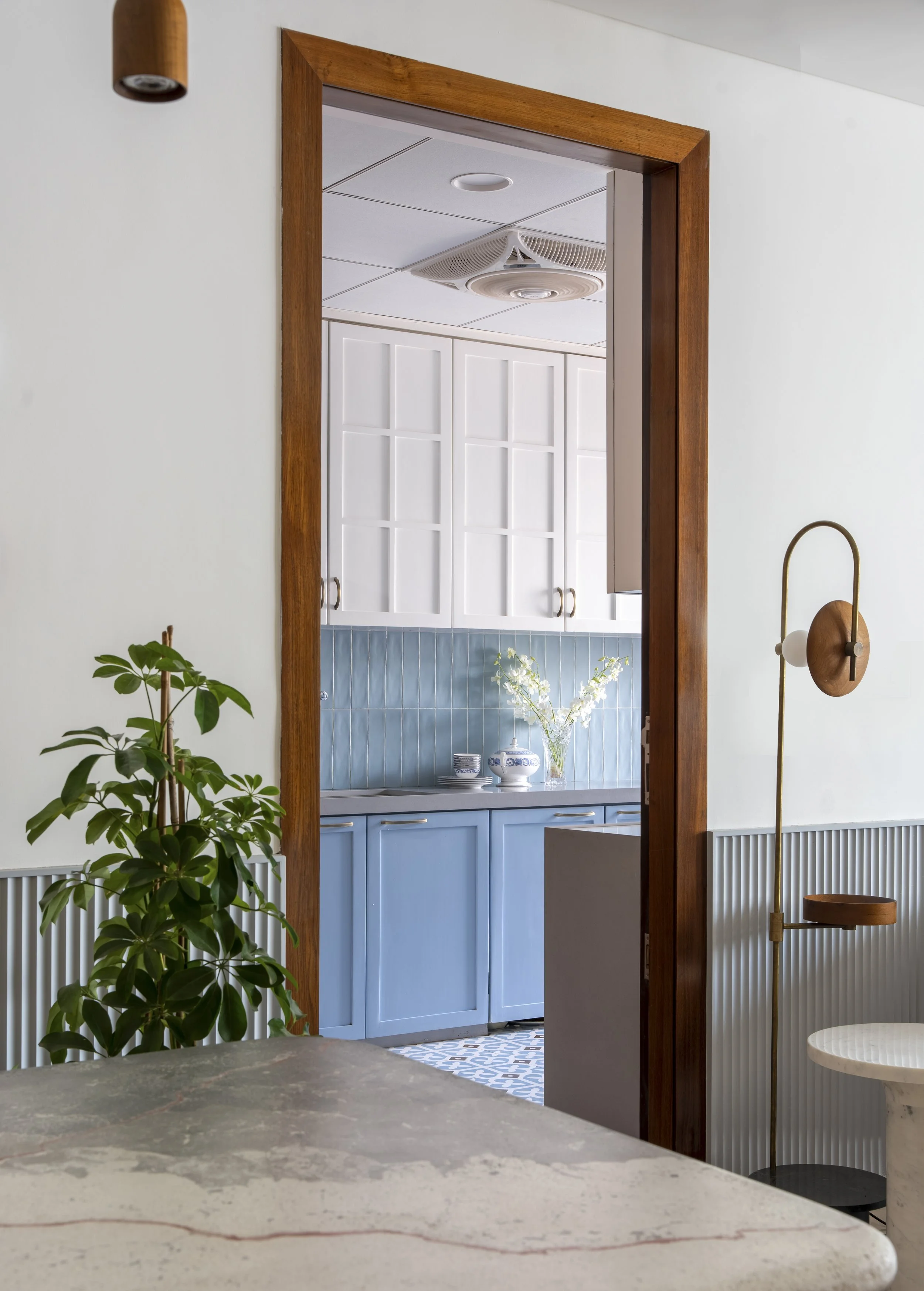 Interior view of a kitchen with white cabinets, blue lower cabinets, and a blue tiled backsplash. A wooden framed doorway leads into the kitchen, with a potted plant and a modern light fixture visible in the foreground.