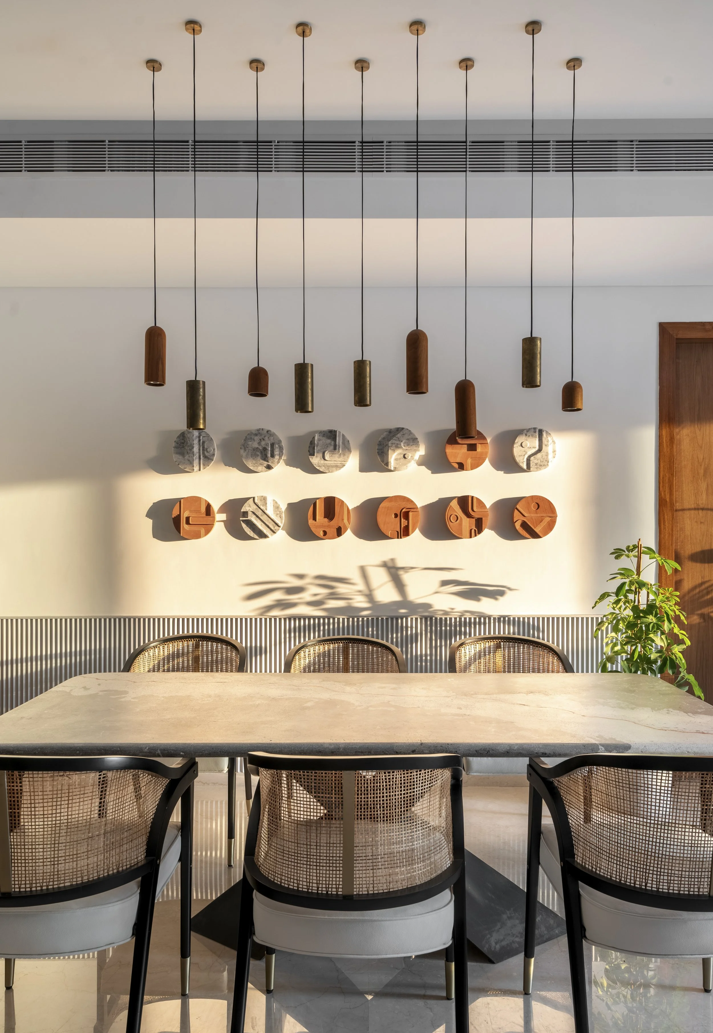 Modern dining room with hanging pendant lights, a long table with wicker-backed chairs, and abstract wall art. Sunlight streams in, creating shadows on the white wall, and there's a potted plant in the corner.