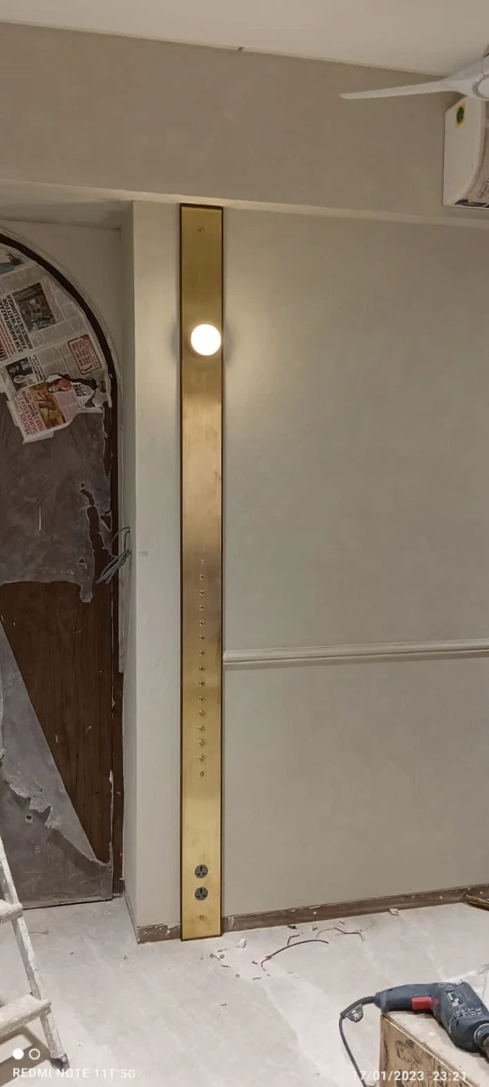 Room with unfinished wall, an arched wooden door covered with newspaper, vertical brass light fixture with a round bulb, electrical outlets, fan, and power drill on the floor.