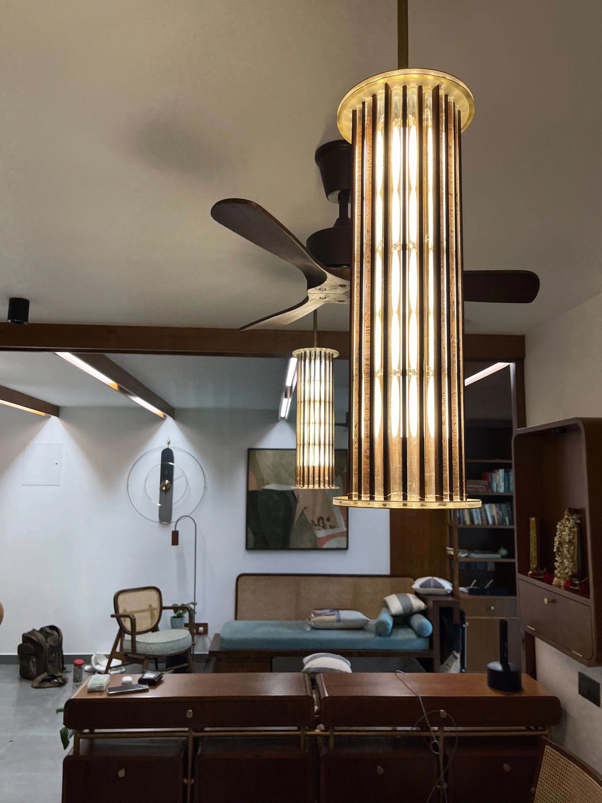 Interior of a modern living room with a large cylindrical pendant light hanging from the ceiling, a ceiling fan with wooden blades, and a lounge area with a couch and cushioned chair. A wall-mounted sculpture and abstract artwork are visible in the b