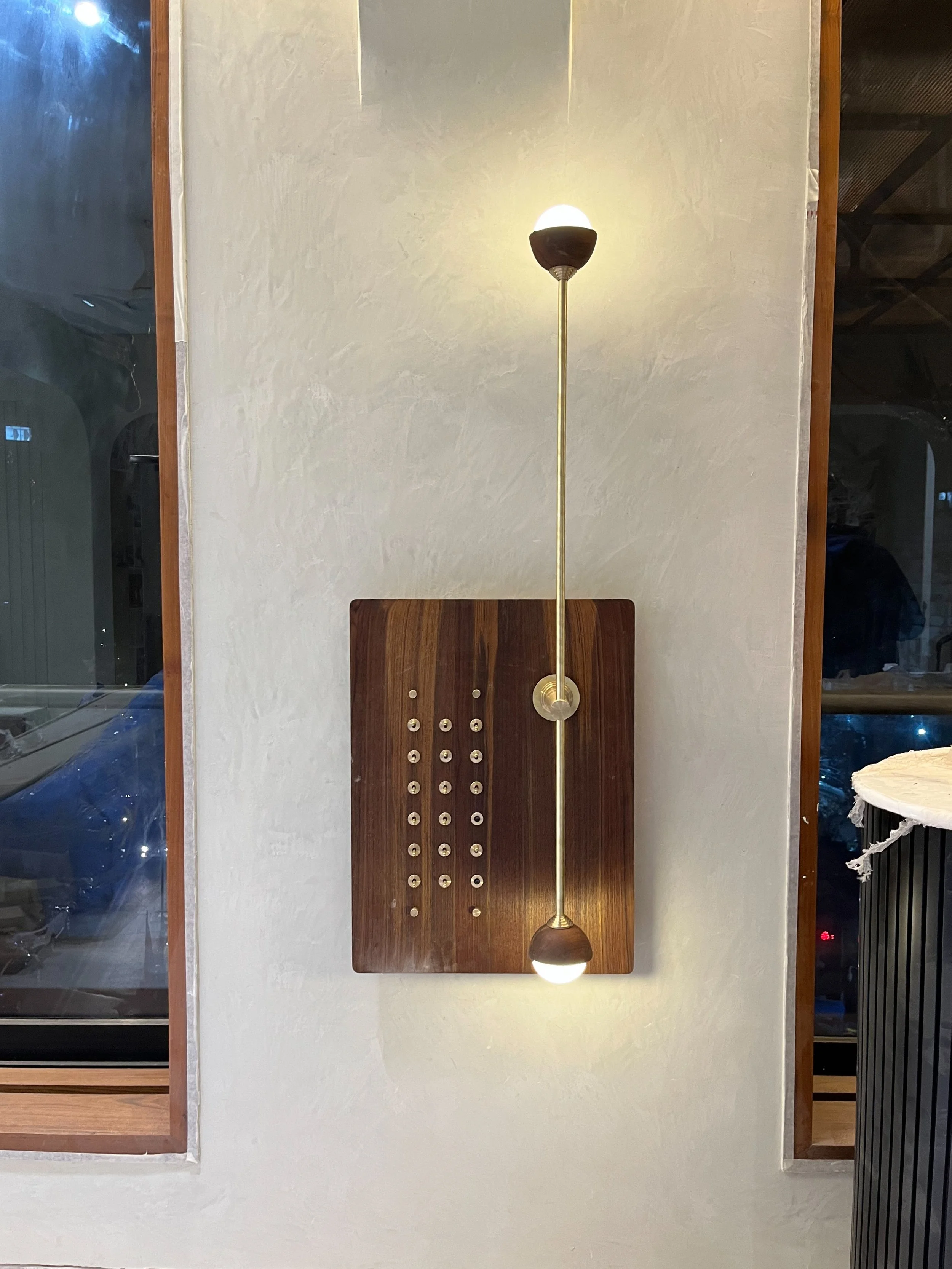 Decorative lighting fixture with dual bulbs and wooden panel on wall near window in modern interior.