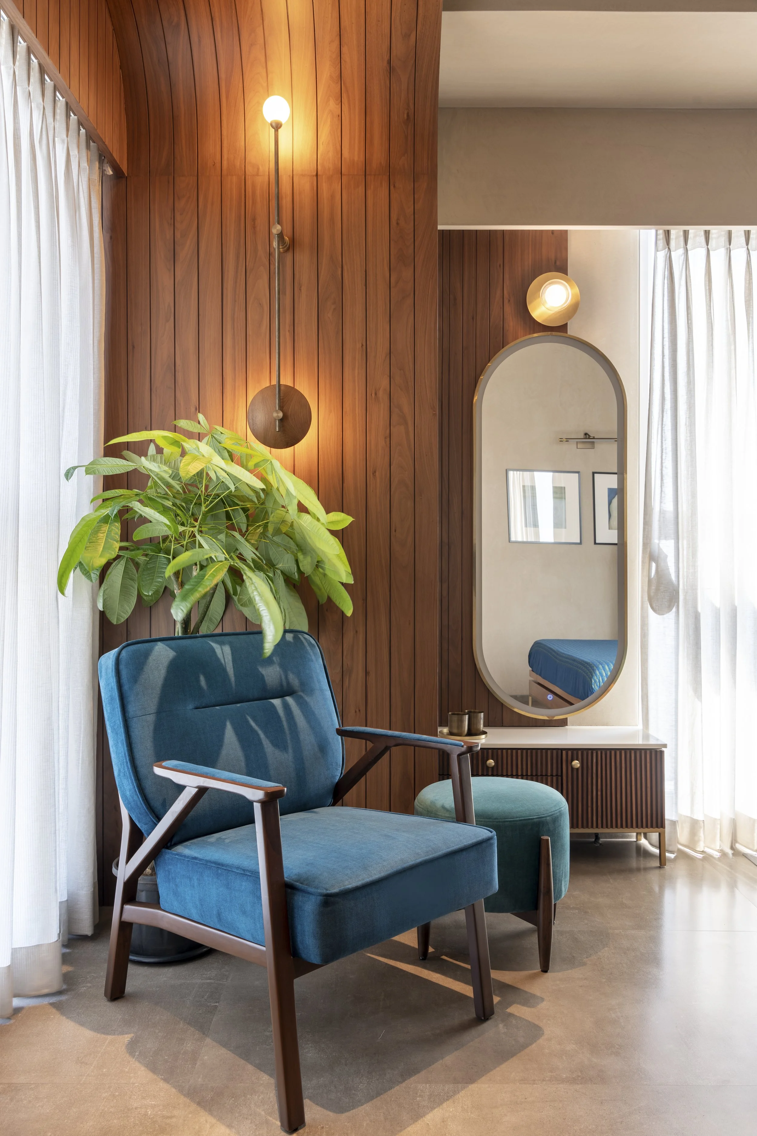 Modern interior space with a blue armchair and ottoman, wooden panel walls, a potted plant, a wall-mounted lamp, and an oval mirror reflecting part of a room. The scene is softly lit with natural light through sheer white curtains.