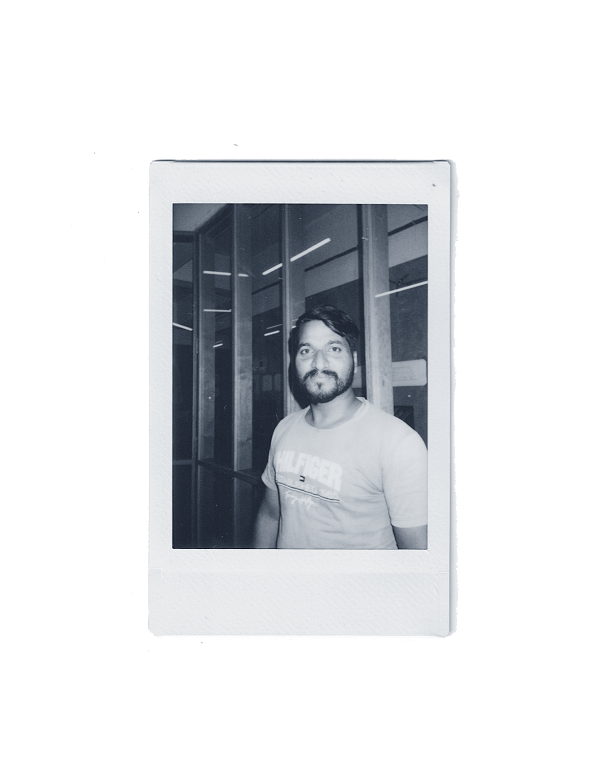 Polaroid photo of a man with a beard and mustache, wearing a T-shirt, standing indoors next to large windows.