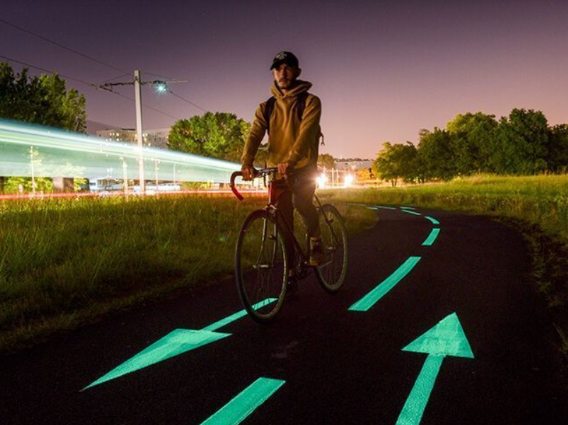 Person riding a bicycle on a glowing bike lane at night, with trees, streetlights, and light streaks in the background.