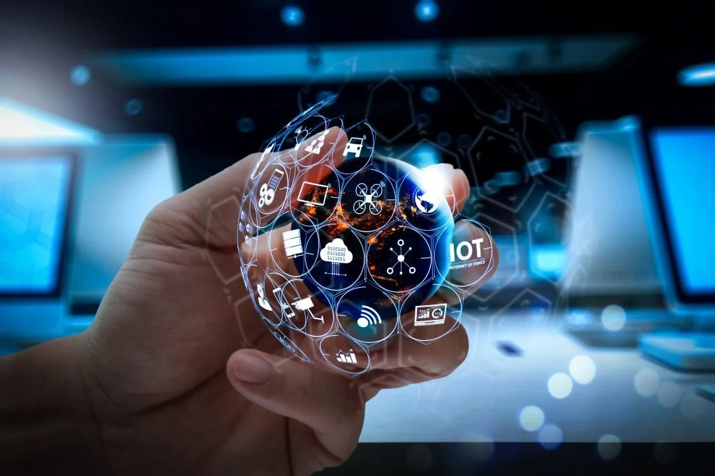 A person holds a holographic globe with icons representing Internet of Things (IoT) technology and connected devices, with computer screens in the background.