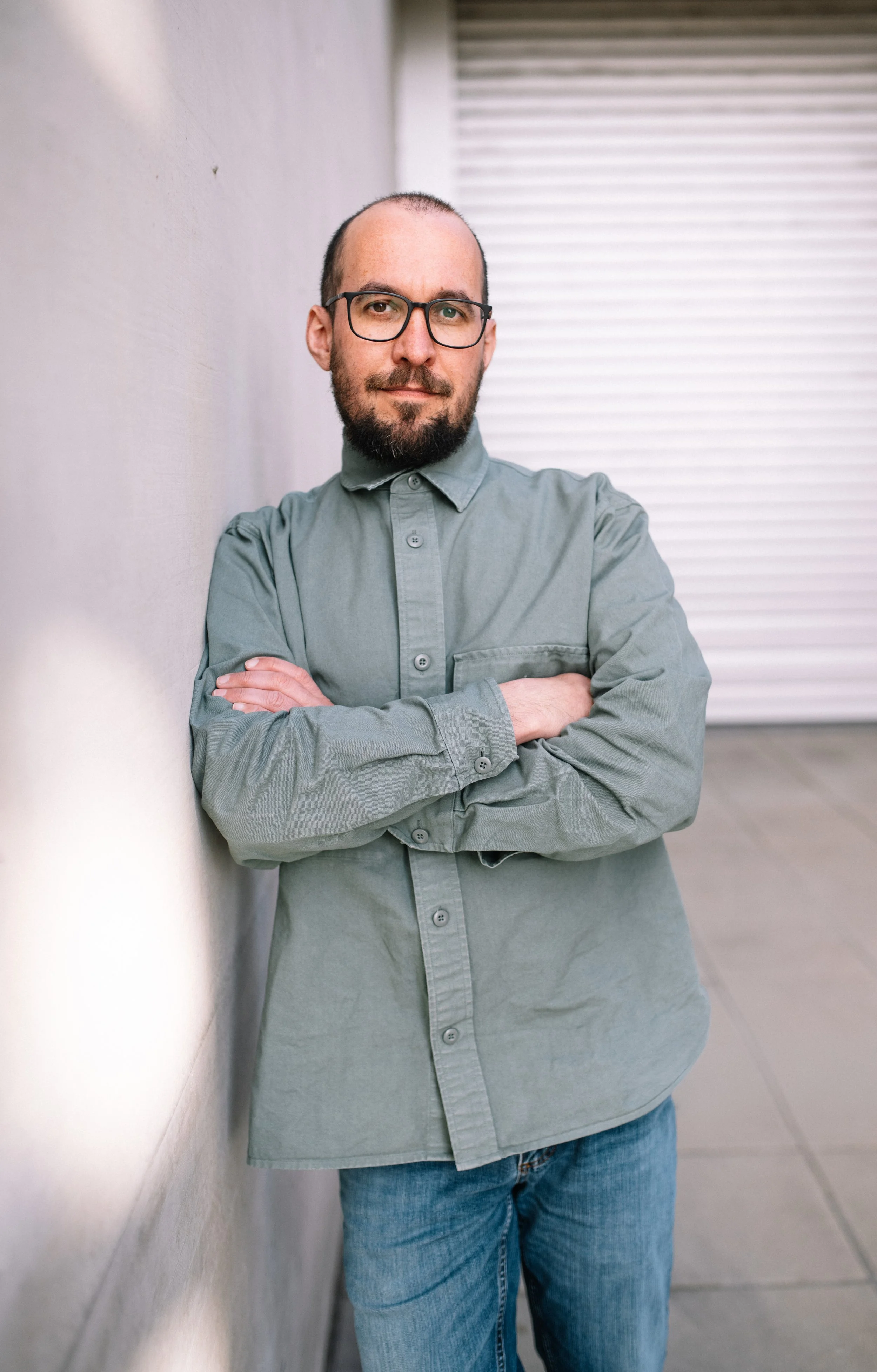 A man with glasses and a beard wearing a gray button-up shirt and blue jeans, standing against a white wall with his arms crossed.