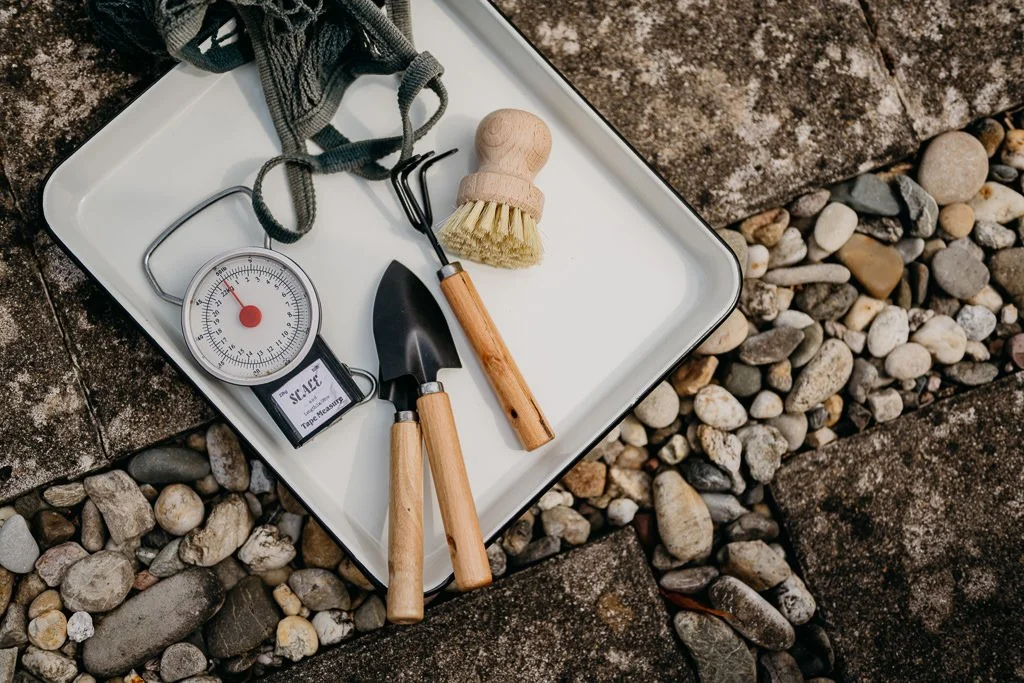 Gardening tools including a small shovel, cultivator, and brush, along with a soil moisture meter on a white tray, placed on a stone and gravel surface.