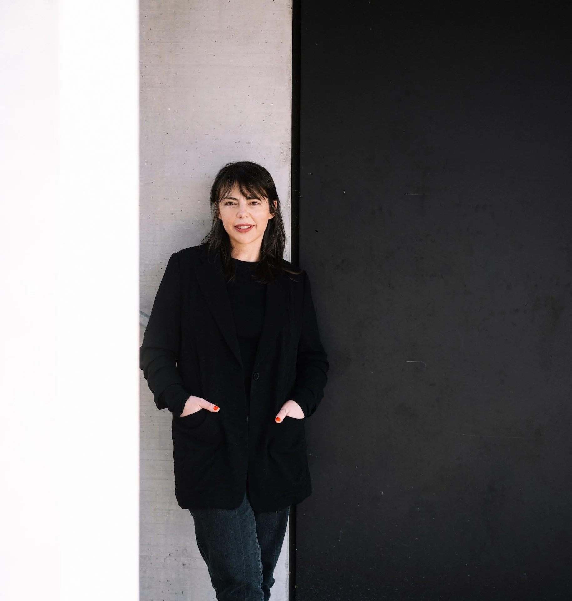 A woman standing against a wall, wearing a black jacket and jeans, with her right arm bent and her hand in her pocket.