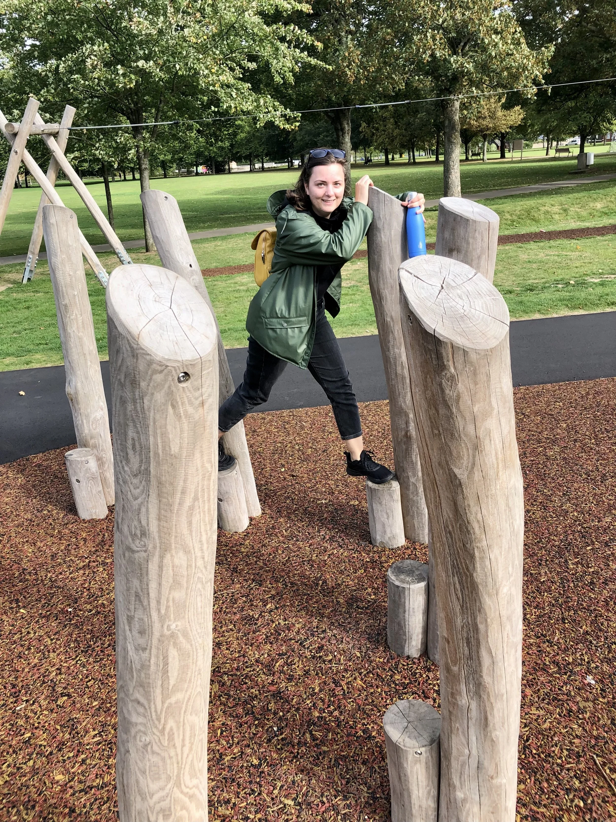 Person balancing on wooden logs in a park playground, surrounded by trees.
