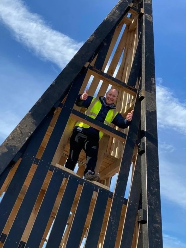 Person in a yellow safety vest standing inside a wooden tower structure, giving thumbs up with a blue sky background.
