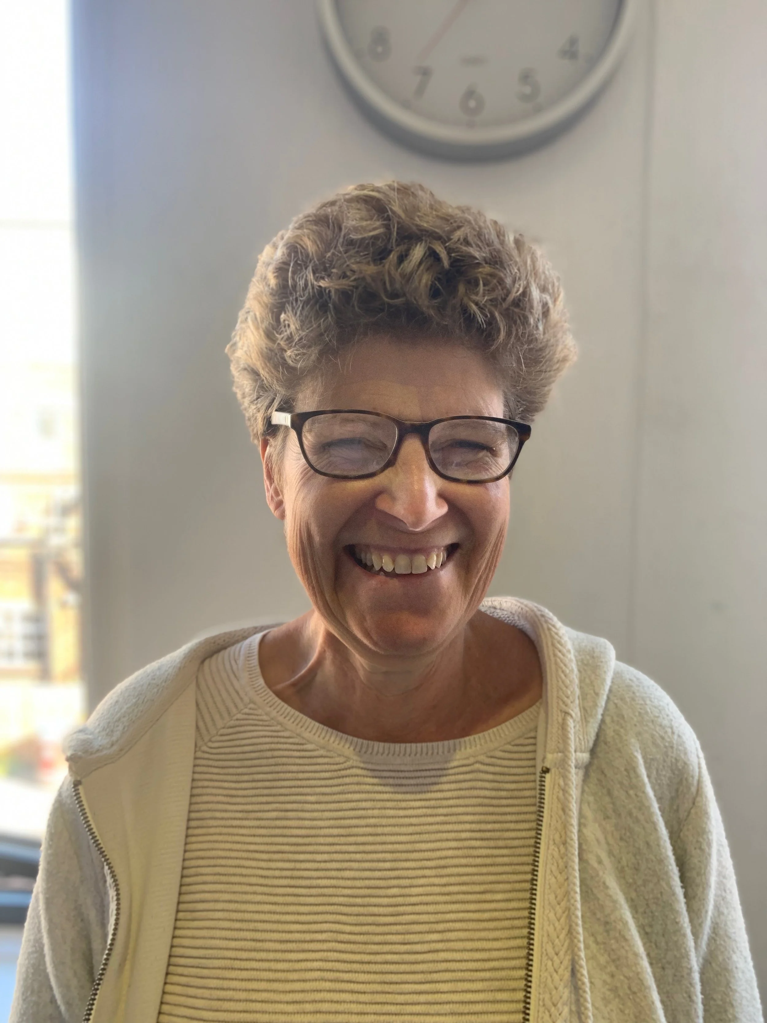 Person wearing glasses and smiling indoors, with a clock on the wall in the background.