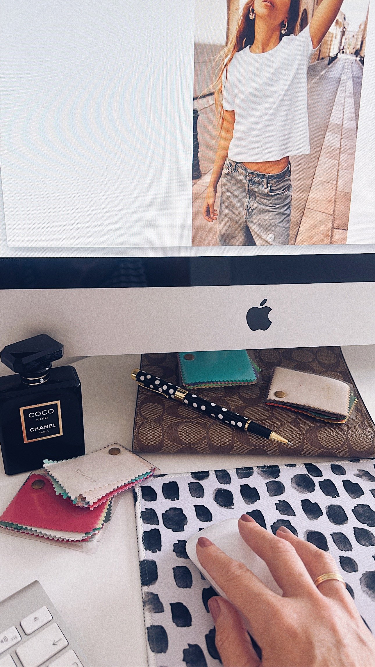 Pantalla de computadora con una foto de una joven con camiseta blanca y pantalones cortos en una calle. En el escritorio hay una botella de perfume Chanel Coco Noir, muestras de telas de diferentes colores, una pluma de bolígrafo y una mano usando una base de limpieza.