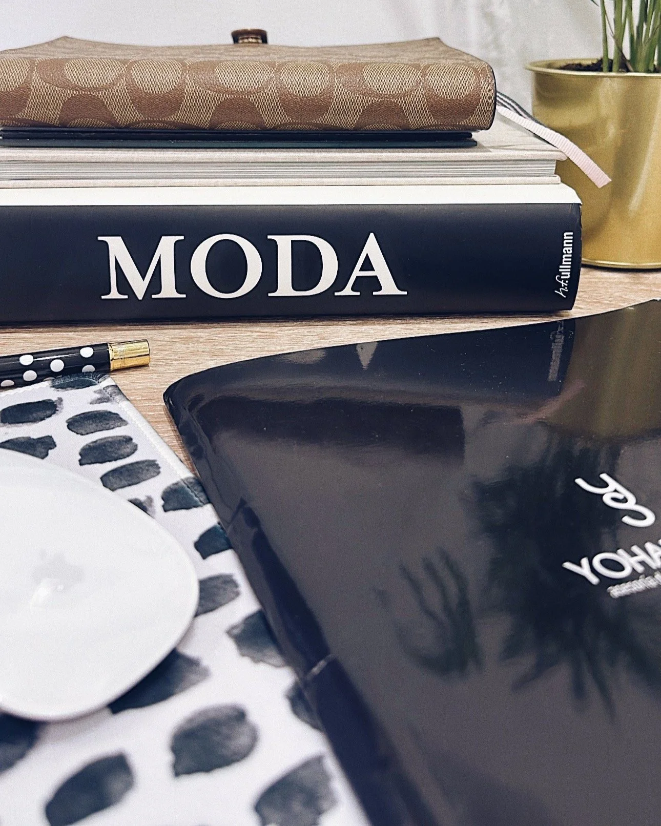 Libros de moda y papelería en una mesa, incluyendo un libro grande con la palabra 'MODA' en la portada, además de una hoja con estampado de puntos y un cuaderno negro con escritura en blanco.