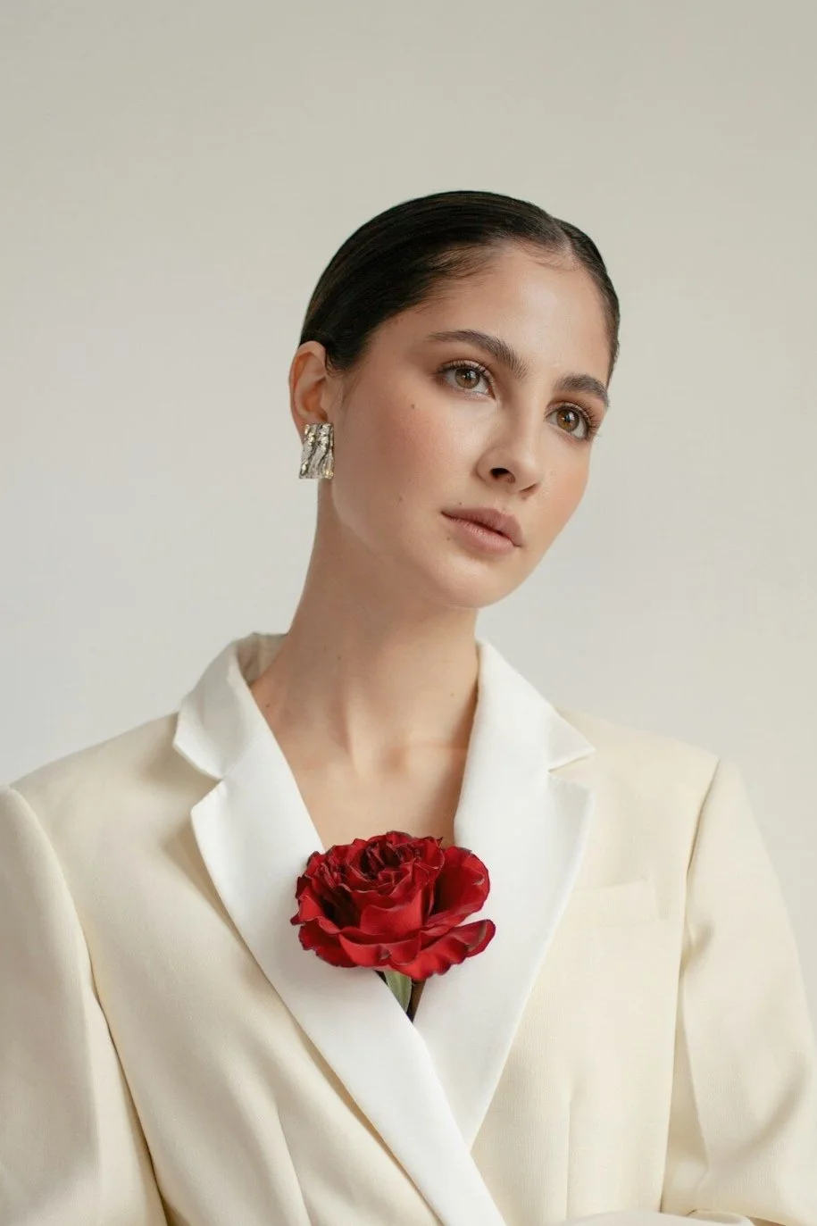 Retrato de una mujer con ropa blanca, orejas plateadas, y sosteniendo una rosa roja en el pecho, contra un fondo claro.