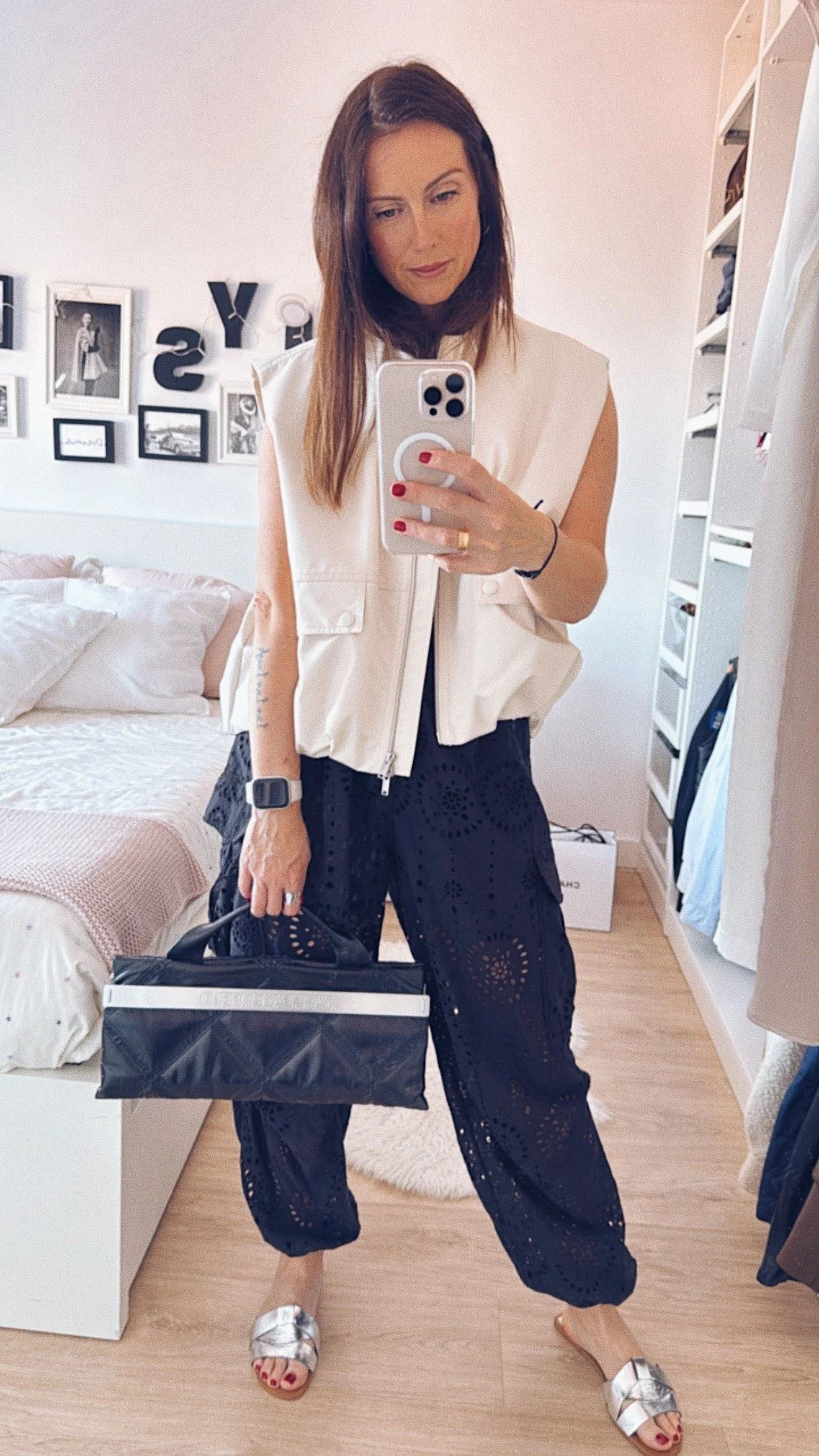 Mujer joven con cabello castaño oscuro, vestida con blusa sans sleeves beige, pantalones negros de diseño perforado y sandalias plateadas, sosteniendo una bolsa negra, tomándose una selfie en un espejo en un cuarto decorado con fotos en la pared y un cabecero de cama en el fondo.