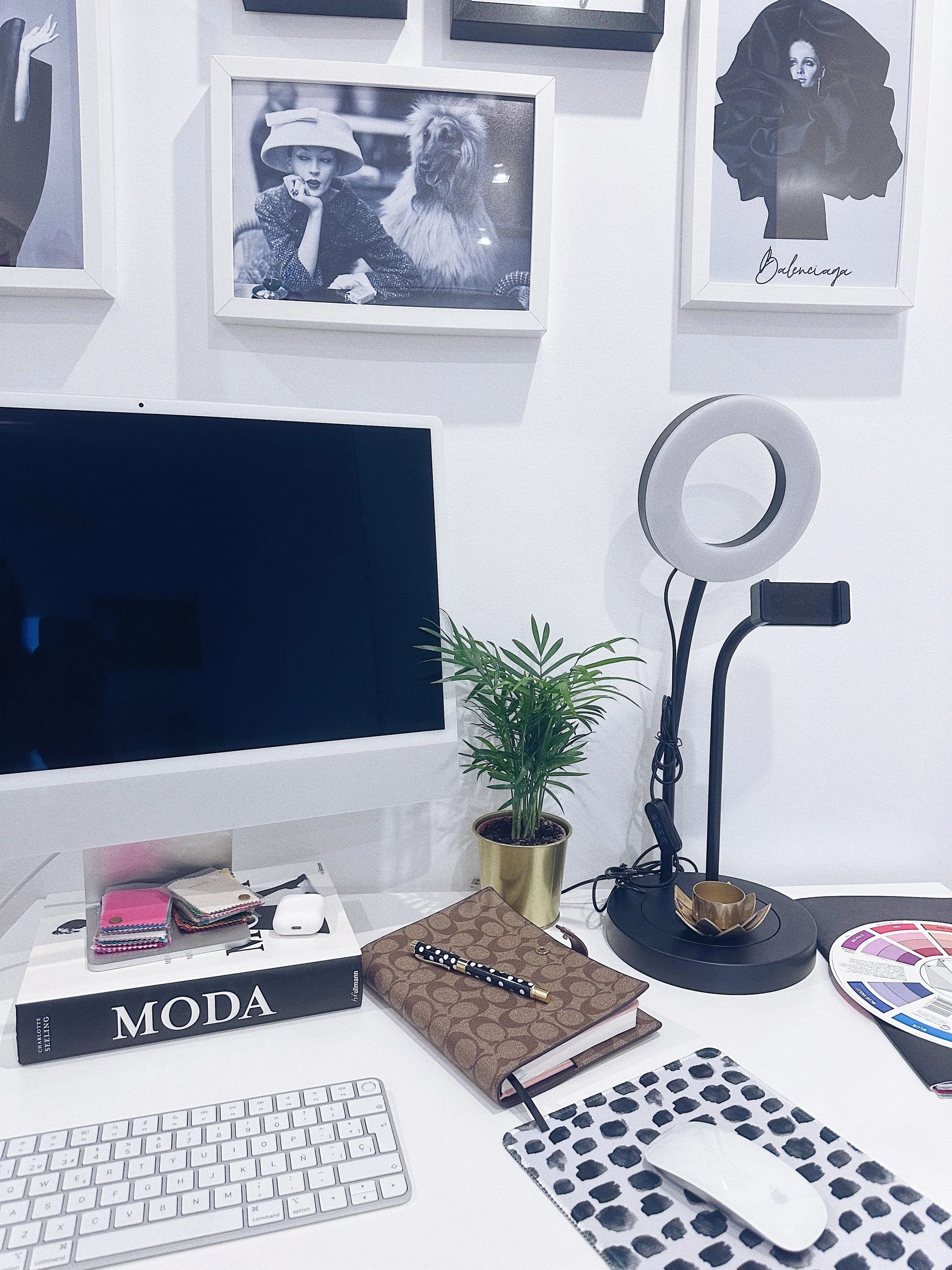 Estación de trabajo con computadora, libro titulado 'MODA', maceta con planta, lámpara de aro y accesorios de oficina. En la pared, cuadros en tonos grises con fotografías y el logo de Balenciaga.