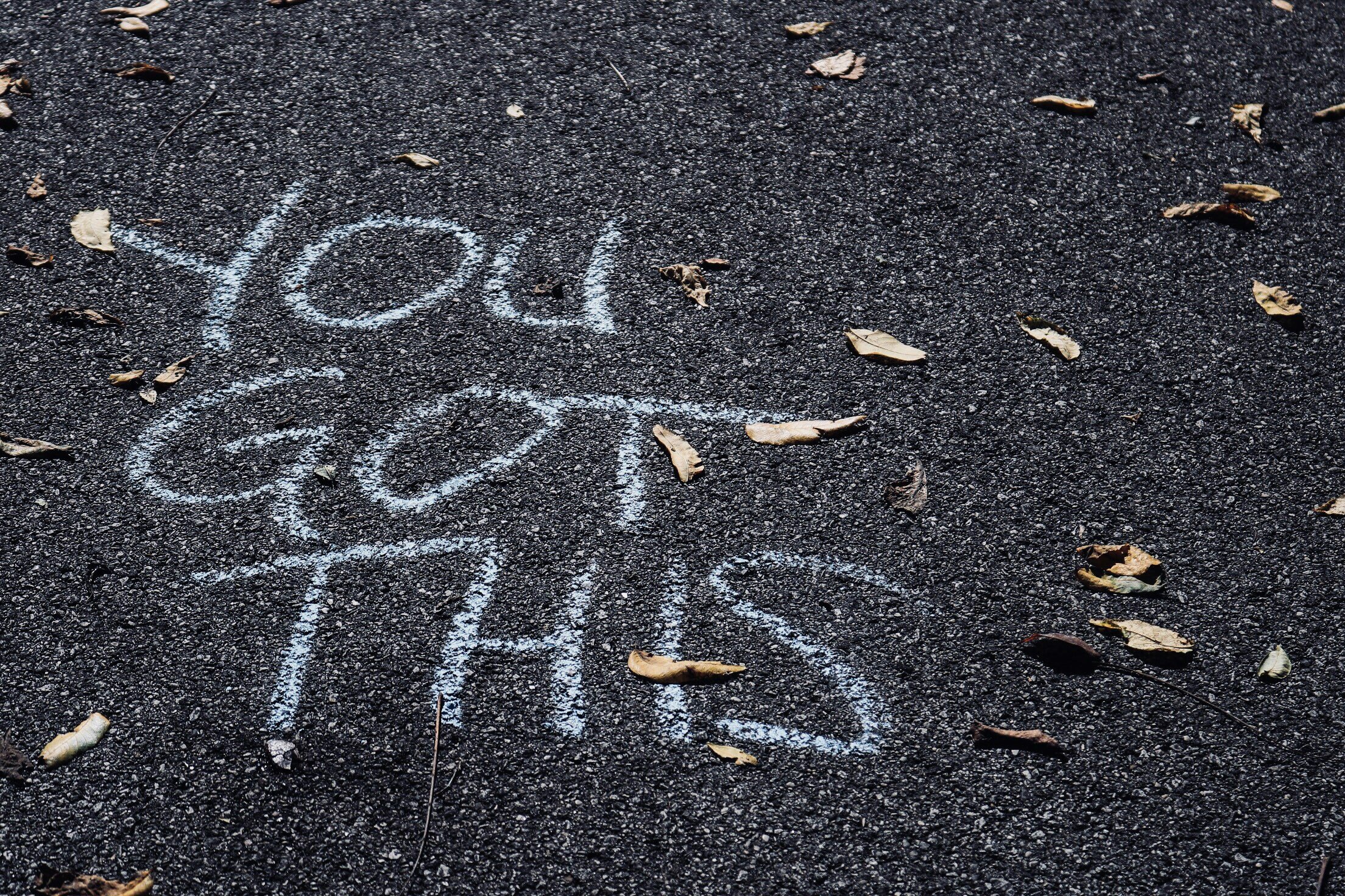 The phrase "You are enough" written with chalk on a dark asphalt surface, surrounded by scattered dry leaves.
