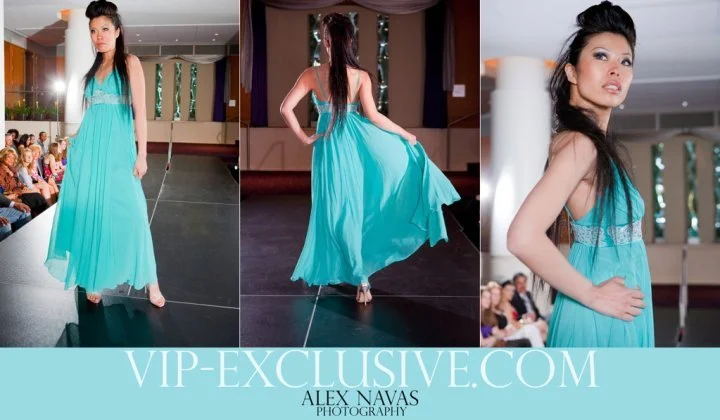 A model walking and posing on a runway during a fashion show, wearing a long turquoise dress with a lace bodice and flowing skirt. Audience members are seated along the runway, and the setting is an indoor event space.