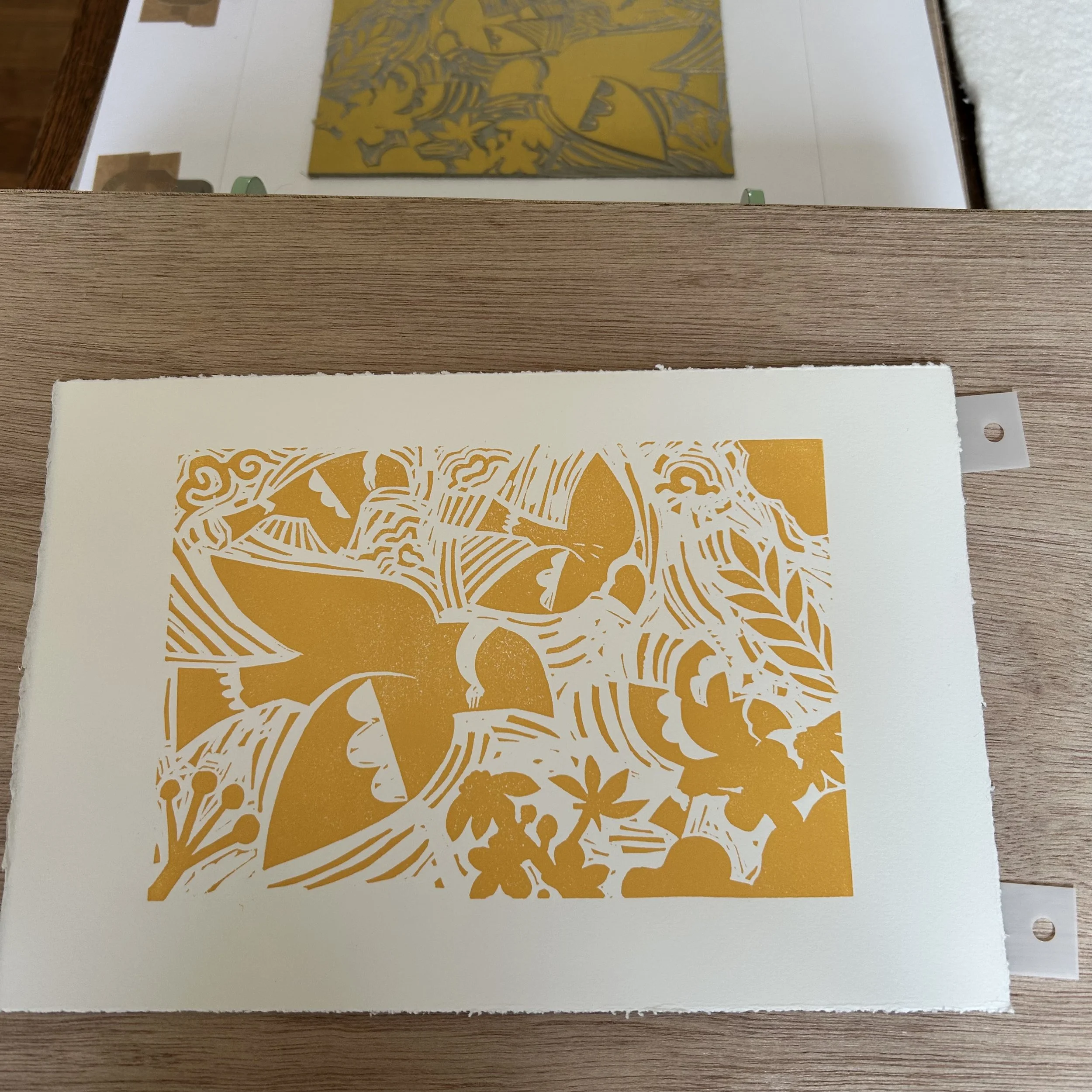 Close-up of a printed paper with an intricate orange and white design of animals and patterns, partially visible on a wooden surface with a mirror reflecting a colorful abstract painting.