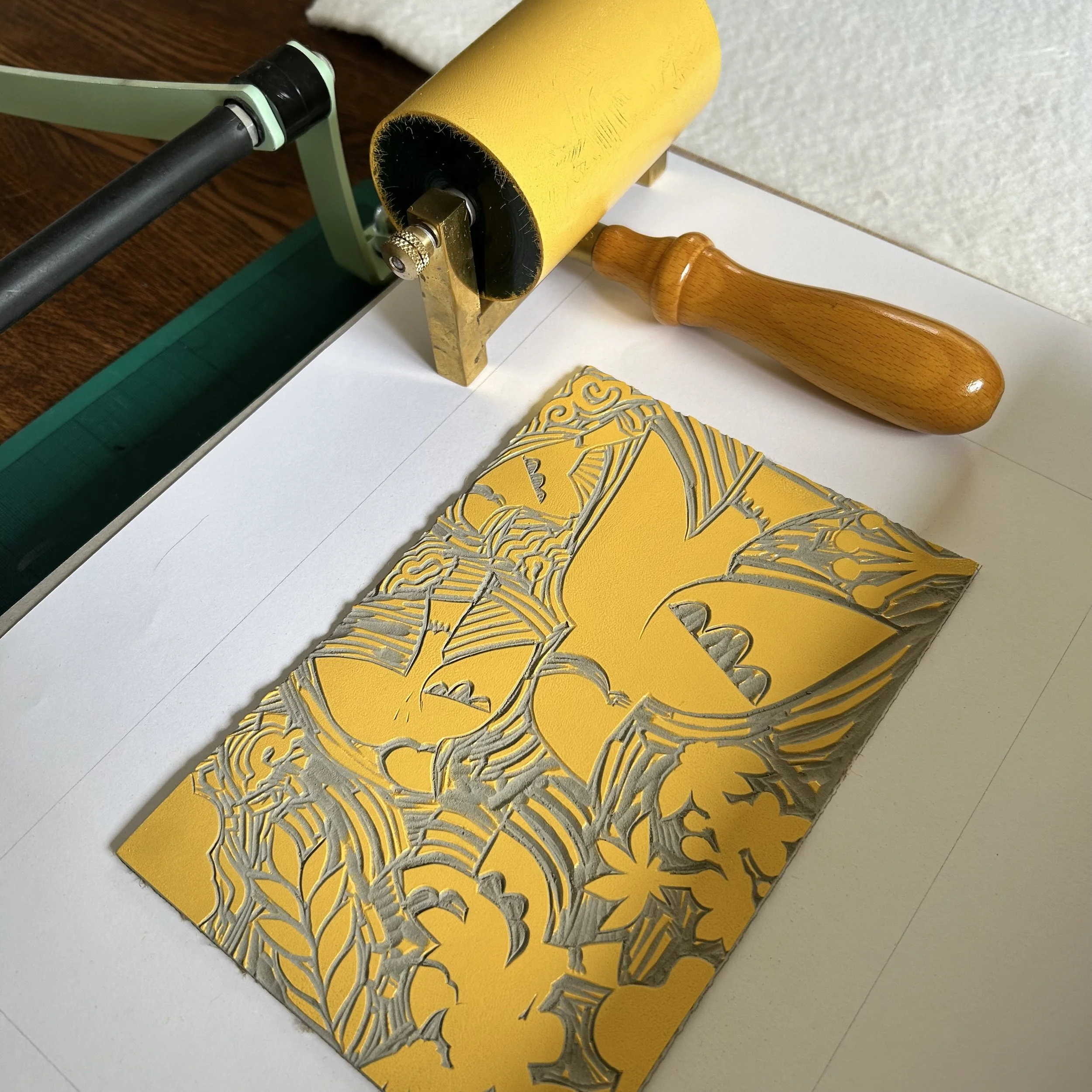 Close-up of a yellow paper with a cut-out leaf design, a foam roller, a quilting ruler, and a small floral patterned fabric on a white surface.