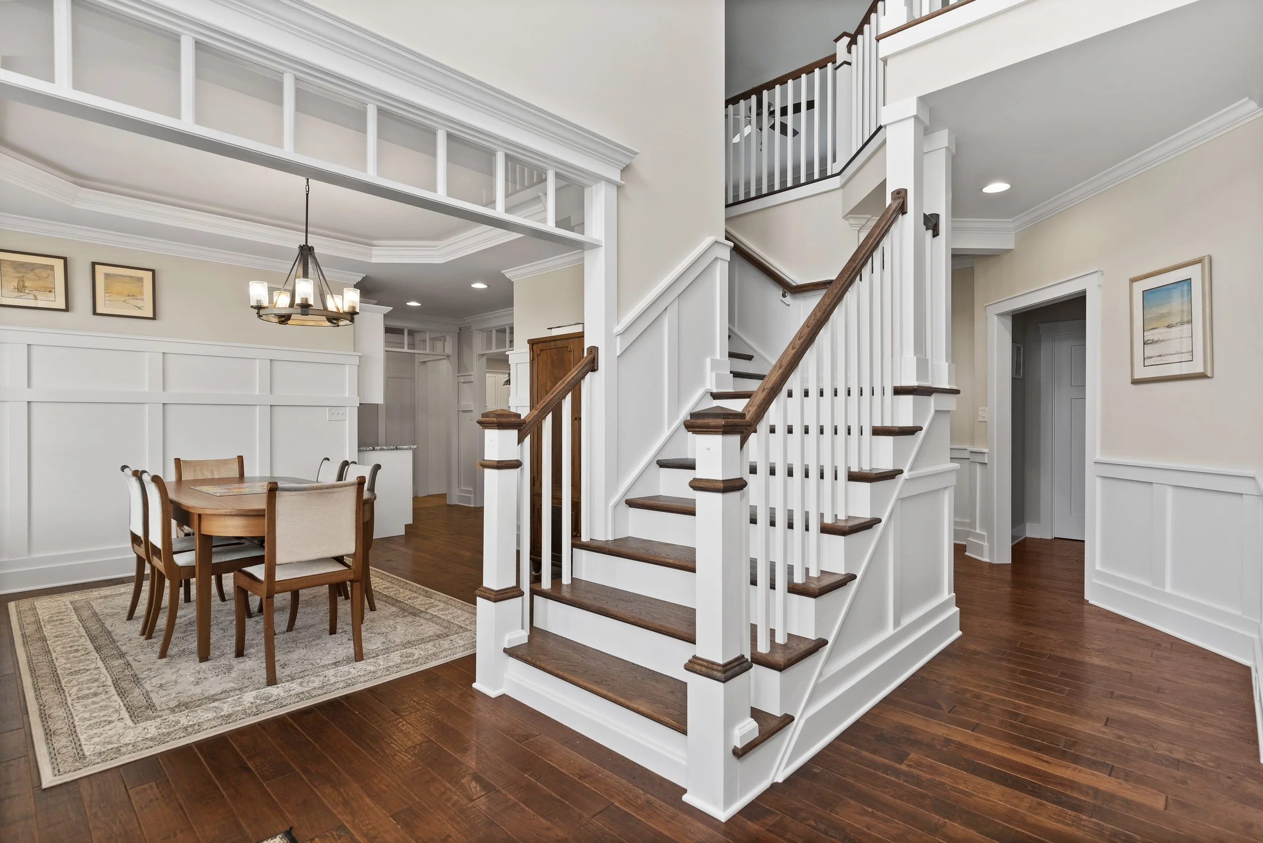 Interior view of a modern home with a wooden staircase, white paneling, and a dining area with a wooden table and chairs on a rug. A chandelier hangs above the dining table; artwork adorns the walls.