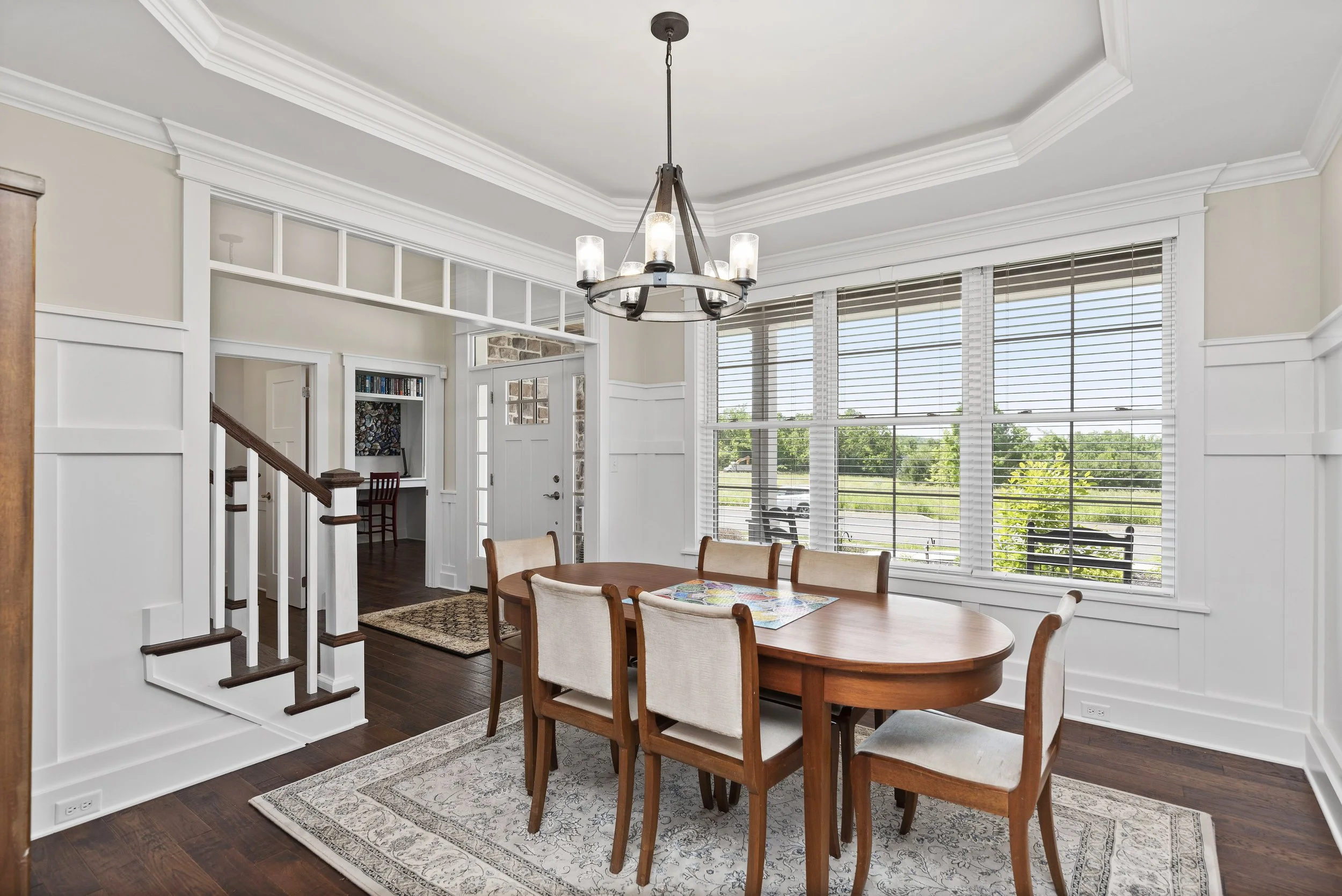 Dining room with wooden table, chairs, chandelier, large windows with blinds, and a staircase in a well-lit home interior.
