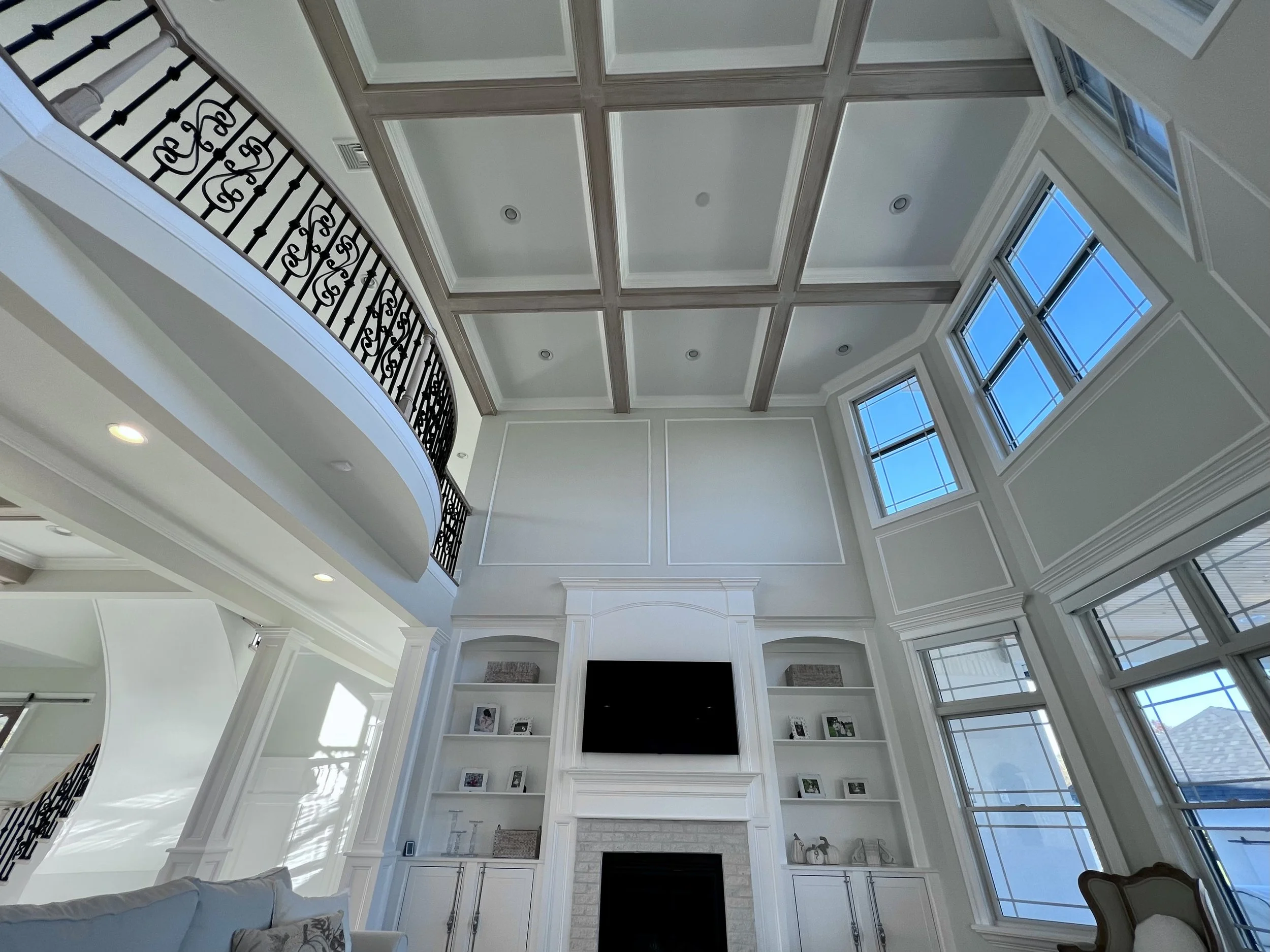 Spacious living room with tall ceiling, coffered design, large windows, iron railings on the upper balcony, fireplace with built-in shelves, modern decor.