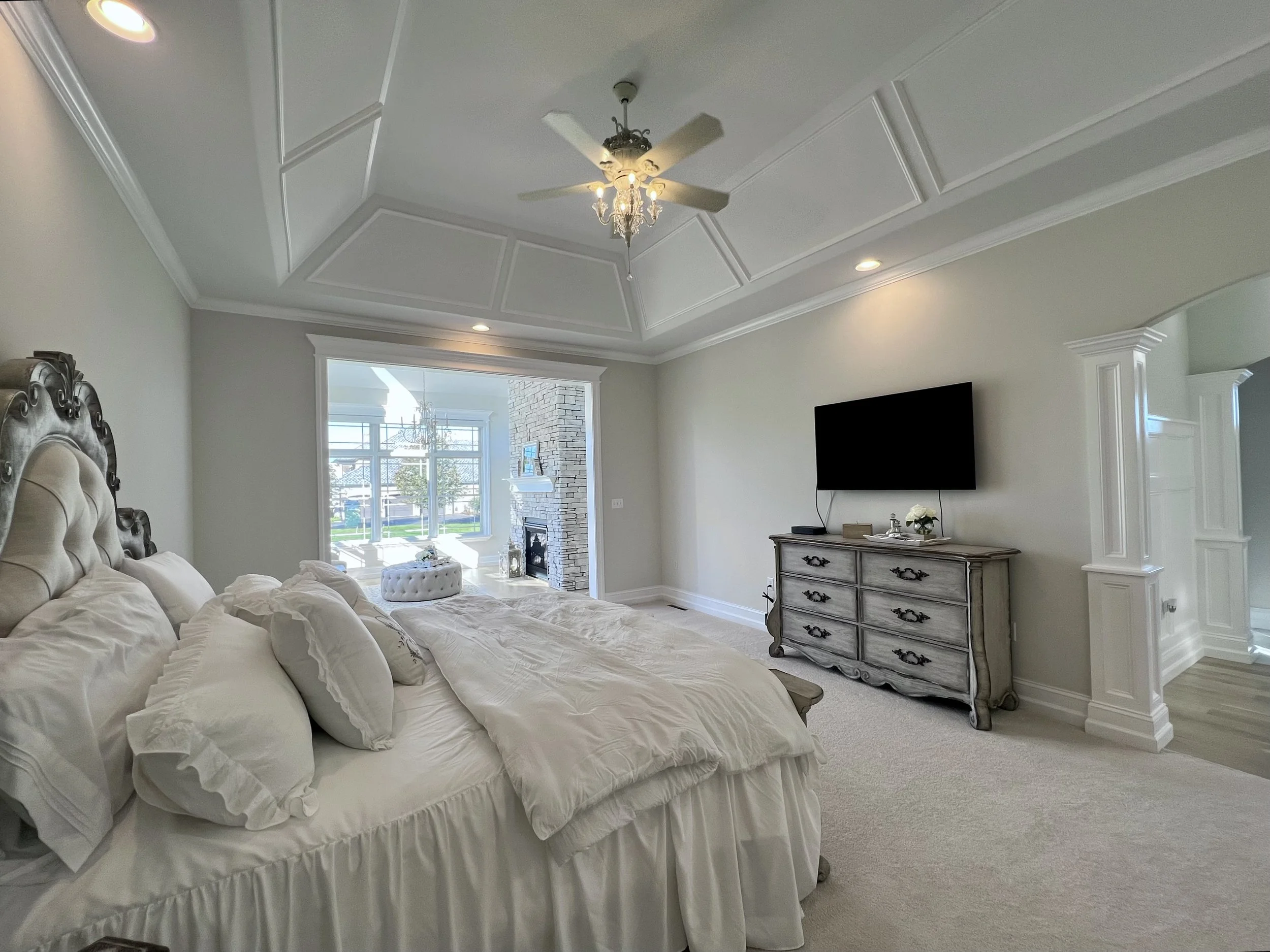 Elegant bedroom with a plush bed, ornate headboard, ceiling fan, flat-screen TV on the wall above a dresser, and an open archway leading to a sunlit sitting area with large windows and a fireplace.