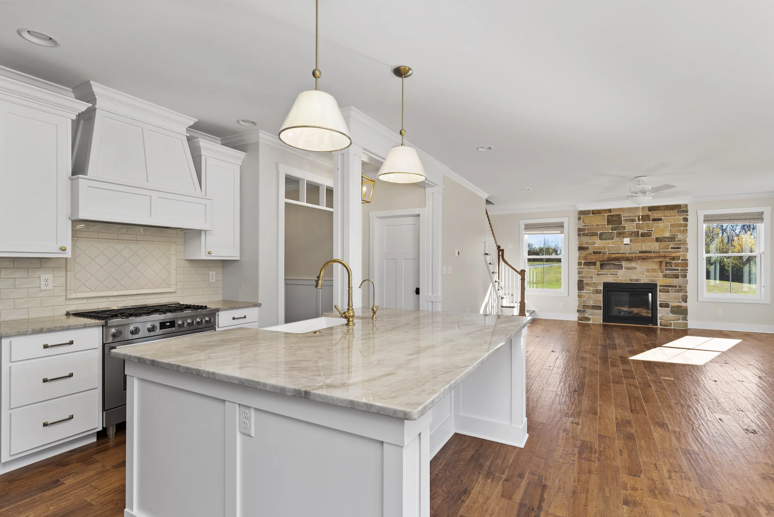 Spacious kitchen and living room with white cabinetry, marble countertops, gas stove, pendant lighting, hardwood floors, stone fireplace, and large windows.
