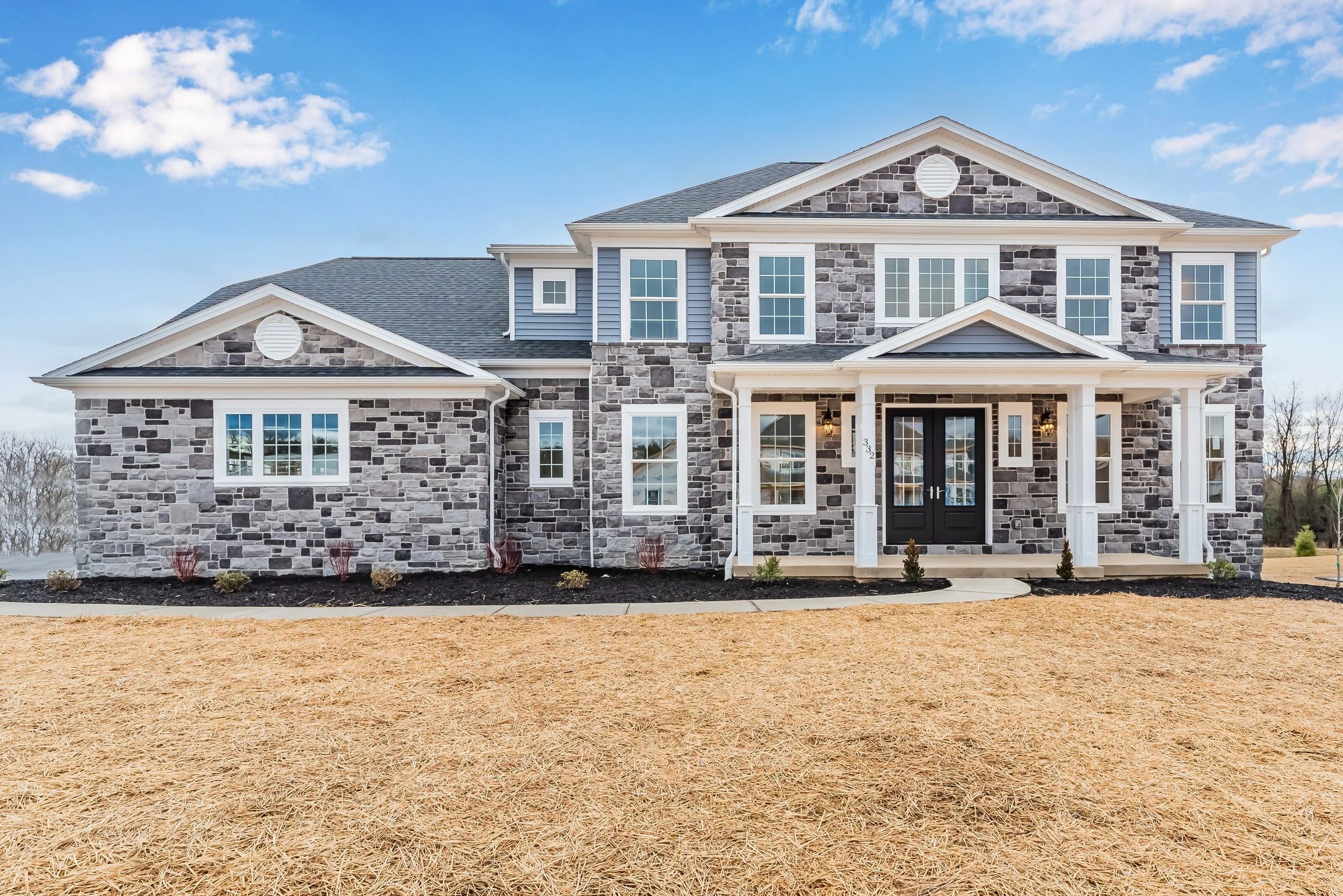 Two-story stone and siding house with front porch and manicured lawn under a blue sky.