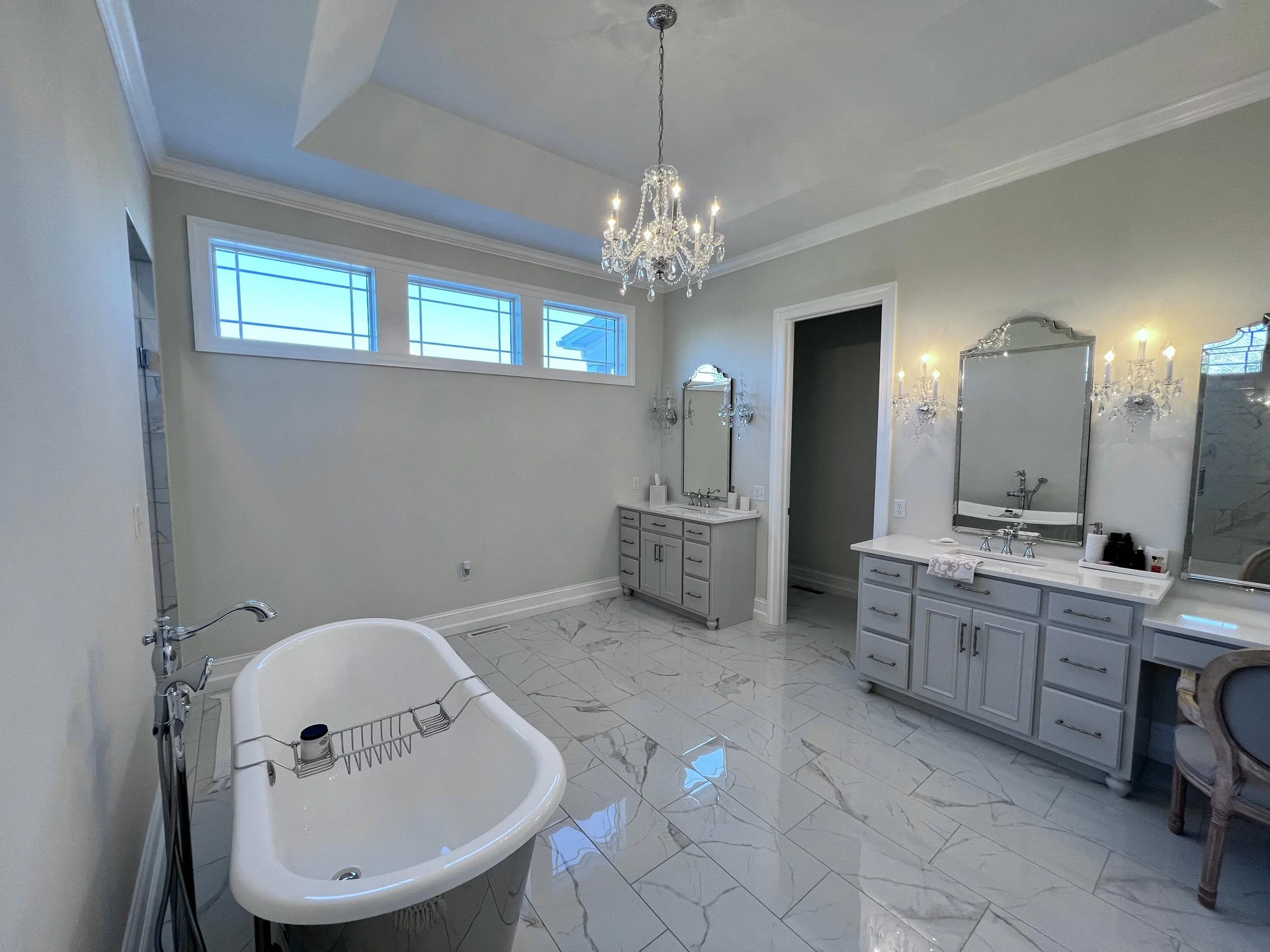 Luxurious bathroom with freestanding tub, gray cabinets, large mirrors, chandelier, and marble tile flooring.